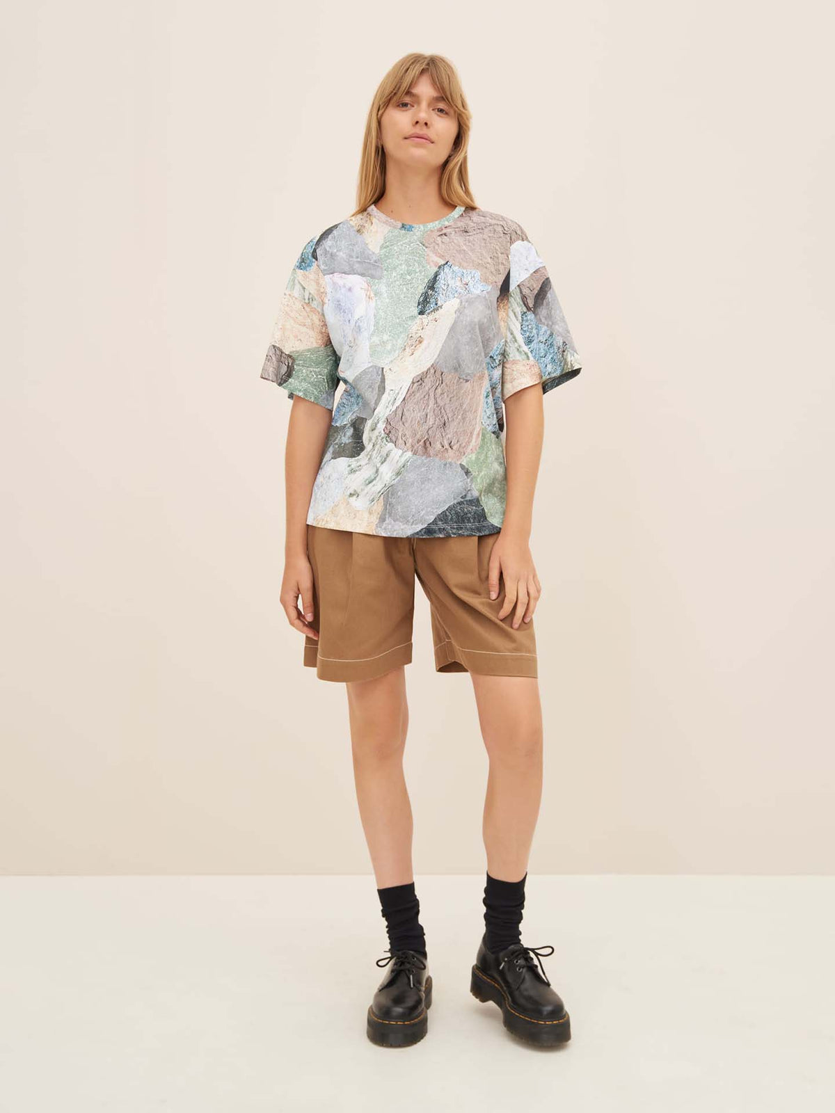 A person stands against a plain background wearing the oversized Mineral Tee by Kowtow, adorned with a colorful abstract pattern, paired with knee-length brown shorts, black socks, and black shoes.