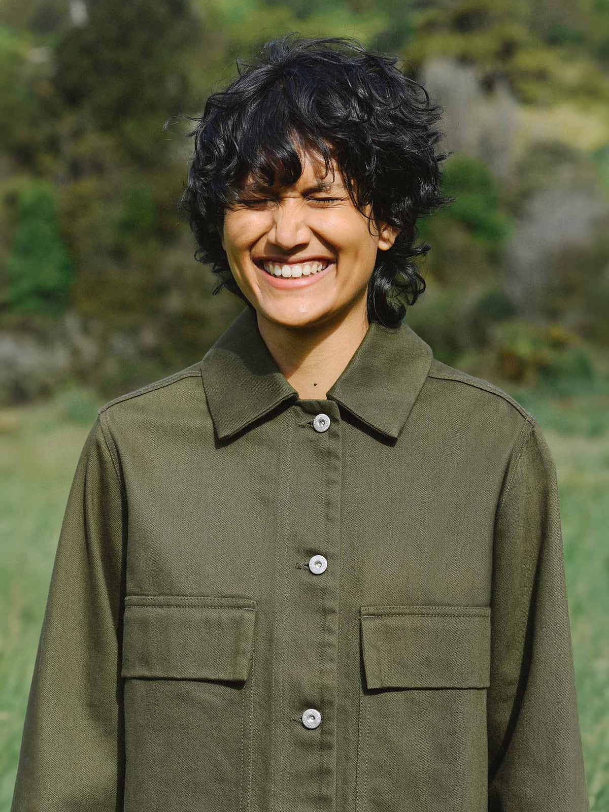 A person with curly black hair smiling outdoors while wearing a Mirror Jacket – Khaki Denim that fits perfectly according to their measurements from Kowtow.