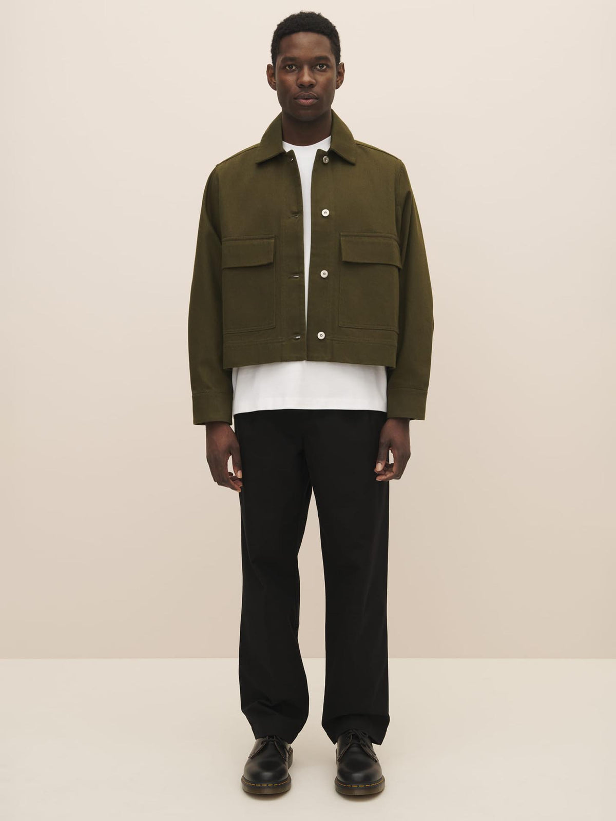 Man standing against a neutral background wearing a Mirror Jacket in Khaki Denim by Kowtow over a white shirt with black trousers and shoes, perfectly matching the size guide for an ideal fit.