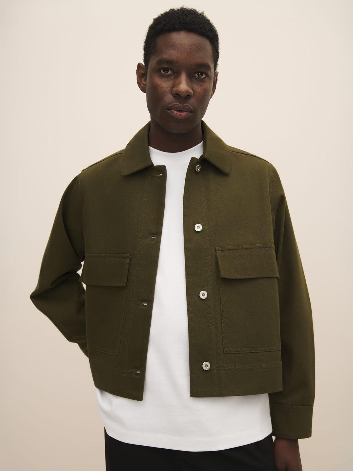 A man wearing a white t-shirt and the Kowtow Mirror Jacket in Khaki Denim with a size guide for the perfect fit, featuring a collar and front pockets.