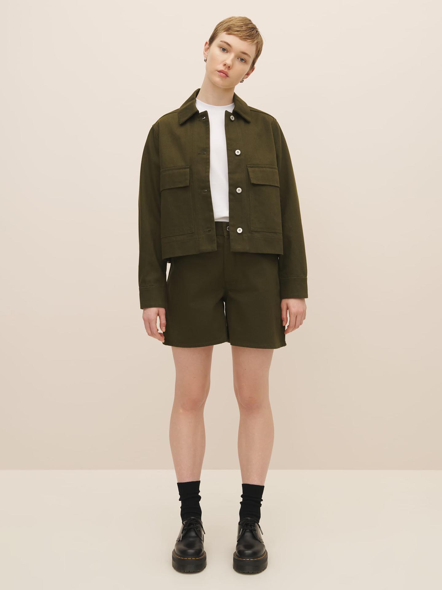 A person posing in a monochrome Mirror Jacket - Khaki Denim ensemble by Kowtow featuring a collared jacket, matching shorts, a white shirt, black socks, and chunky black shoes. For the perfect fit, please consult our size chart.