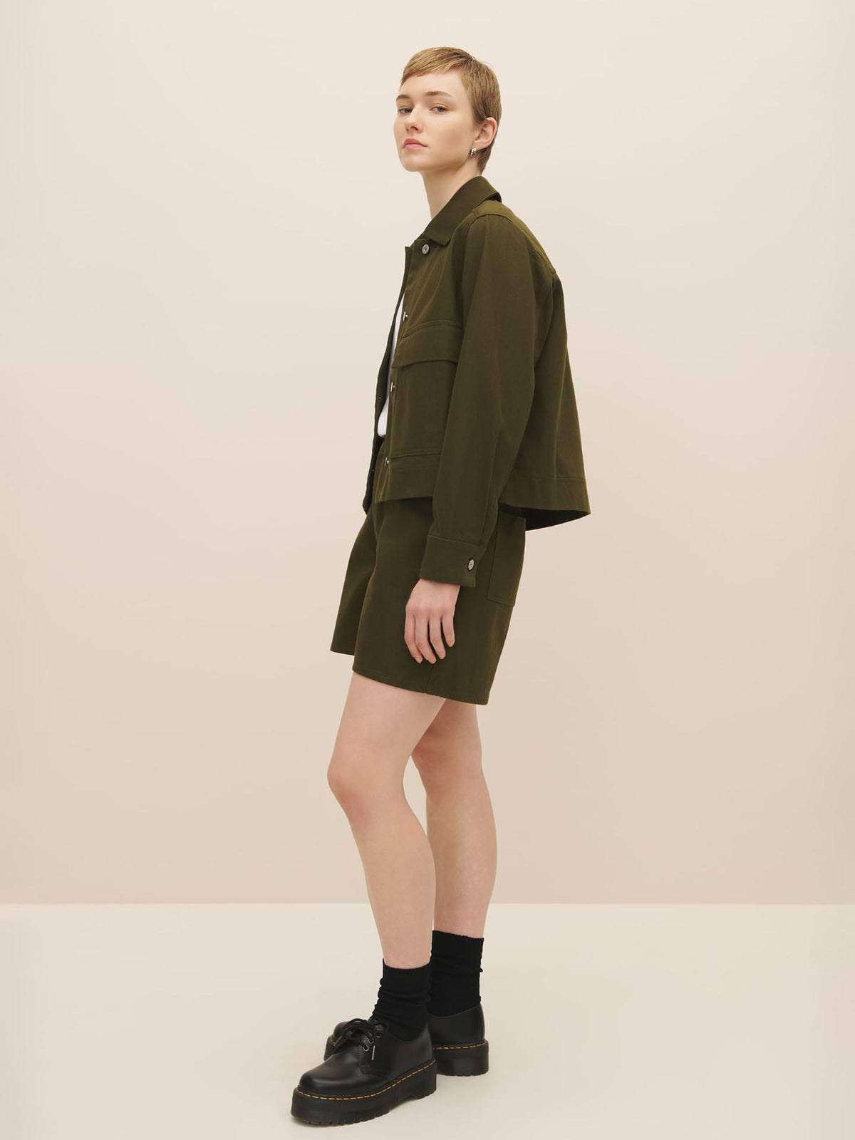 A person standing sideways wearing a Mirror Jacket - Khaki Denim by Kowtow over a white top, matching shorts, black socks, and black platform shoes, all according to the size guide.