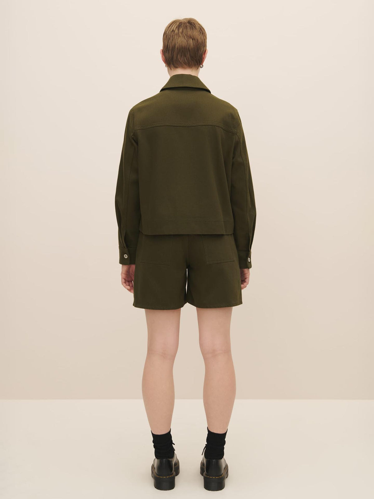 Person standing with their back to the camera, wearing a Kowtow Mirror Jacket in Khaki Denim and matching shorts with black socks and boots. For the perfect fit, refer to our size guide.