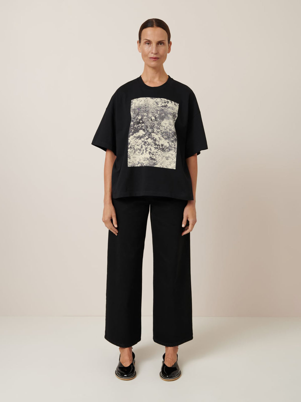 A person poses against a plain background, donning Kowtow&#39;s Monochrome Tee in black with an abstract rectangular print, styled in an oversized fit, paired with wide-leg black pants and coordinating shoes.