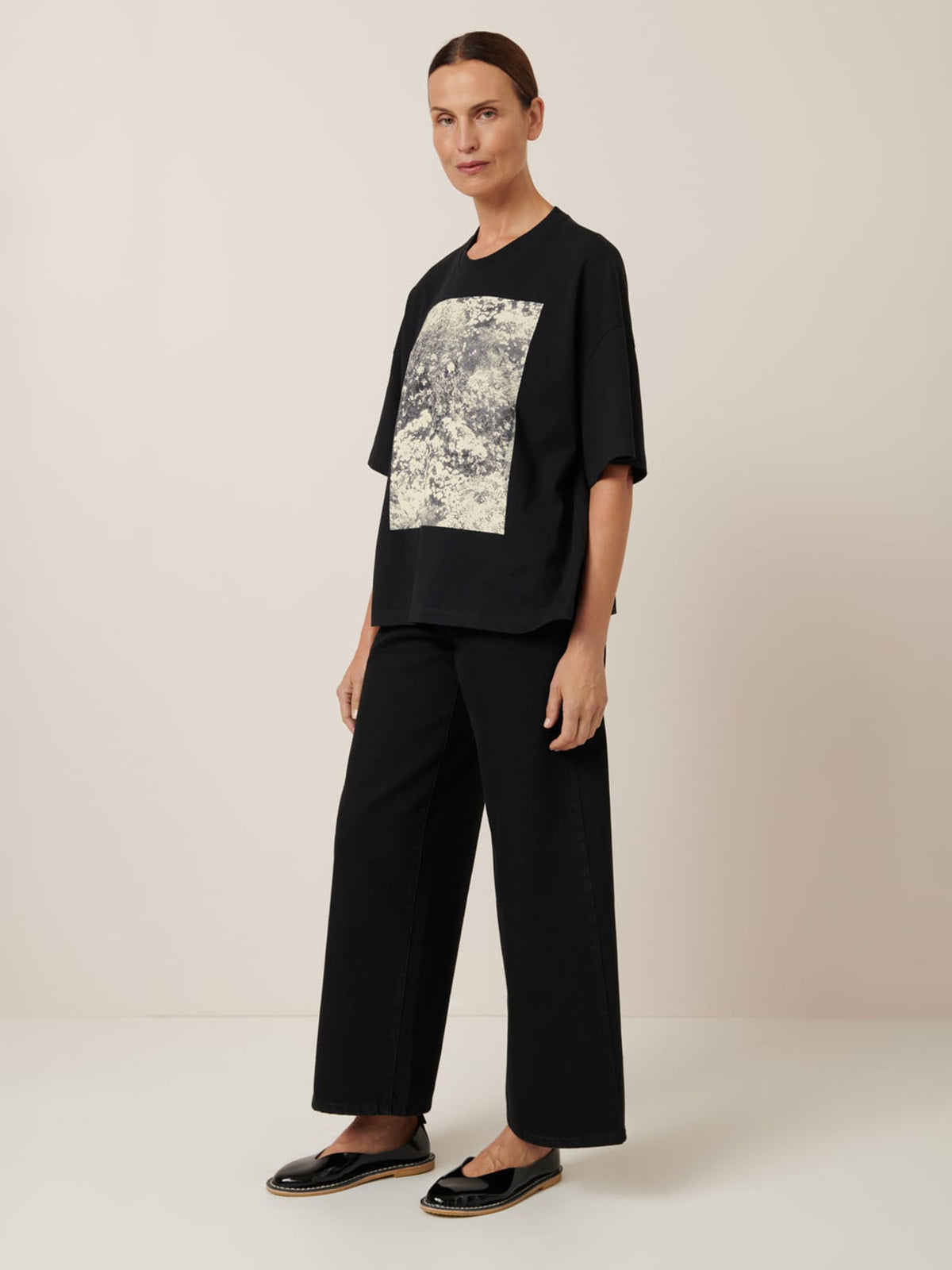 A person stands against a plain background wearing Kowtow&#39;s Monochrome Tee, a black T-shirt with a graphic design, paired with black wide-leg pants that create an effortless oversized fit.