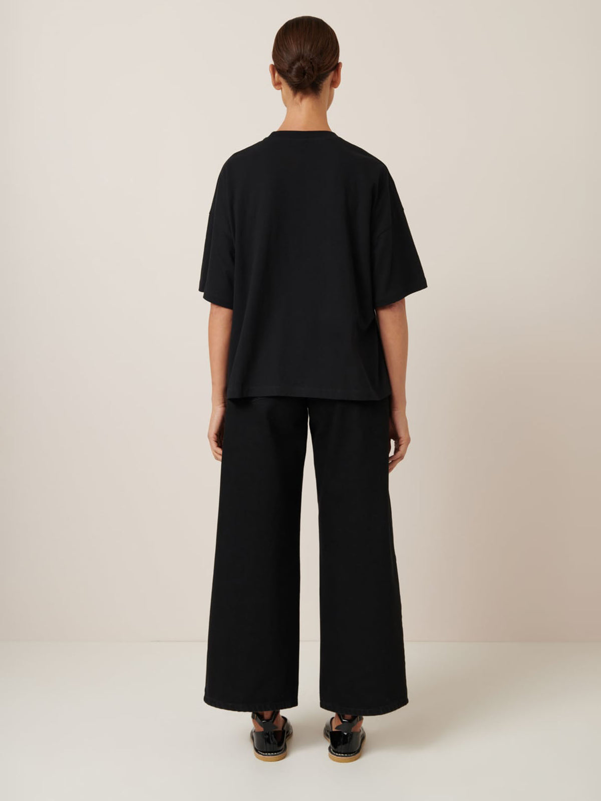 A person with tied-back hair and their back to the camera showcases Kowtow&#39;s Monochrome Tee with an oversized fit, paired with wide pants and sandals against a plain, light-colored background.