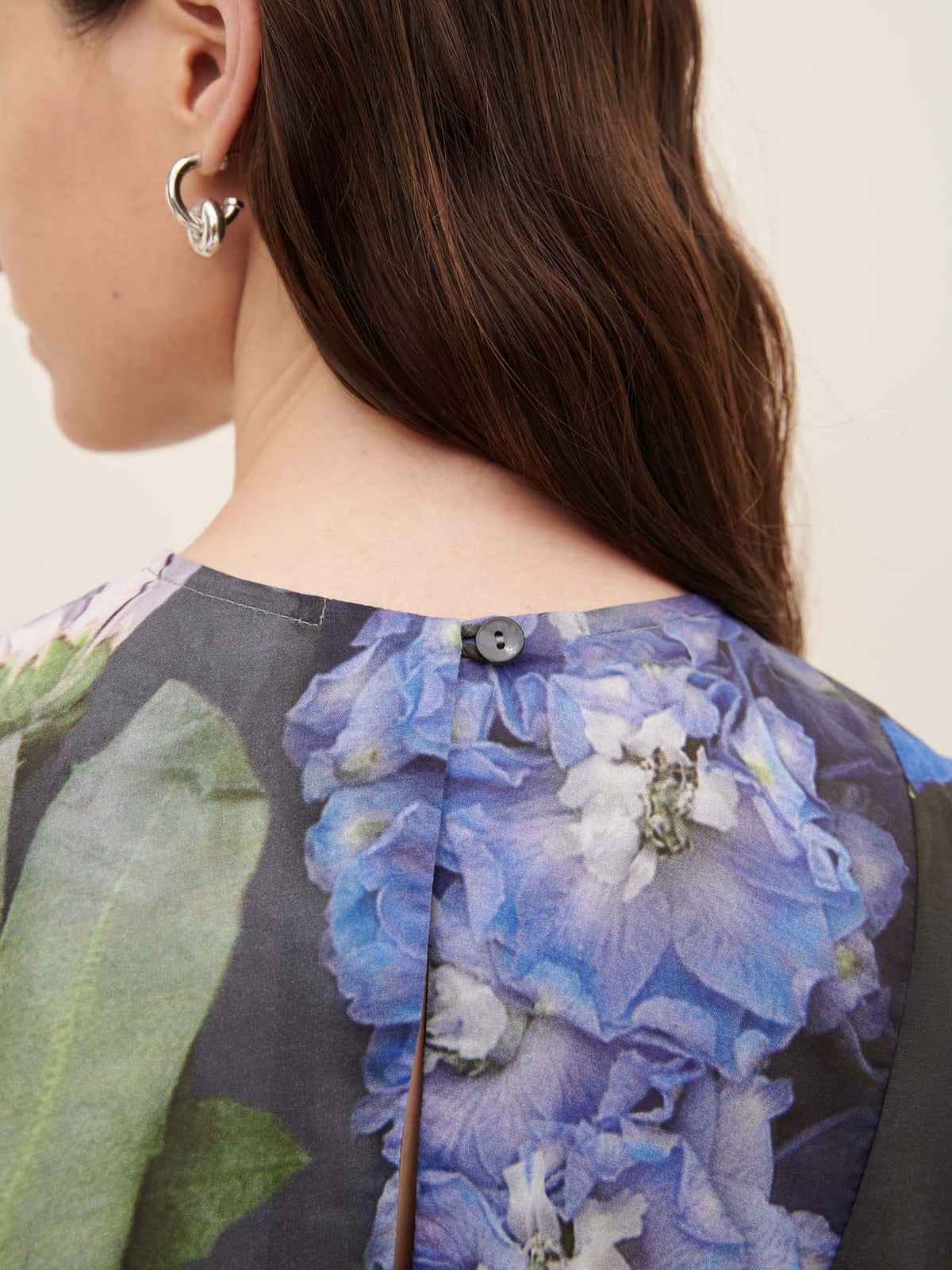 A woman with long brown hair, wearing silver hoop earrings and a Mosaic Dress by Kowtow, featuring a floral pattern in blue and green hues, is viewed from the back, highlighting its oversized fit.