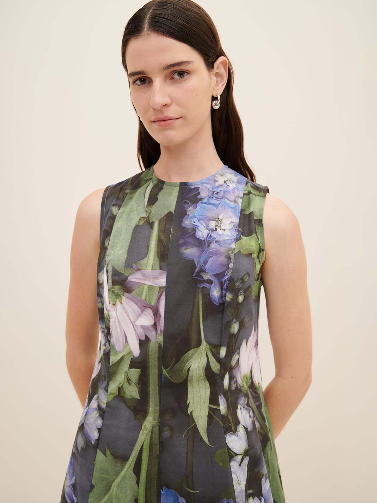 A person with long hair, dressed in the oversized fit Mosaic Dress by Kowtow, adorned with a floral pattern and sleeveless design, stands against a plain background.