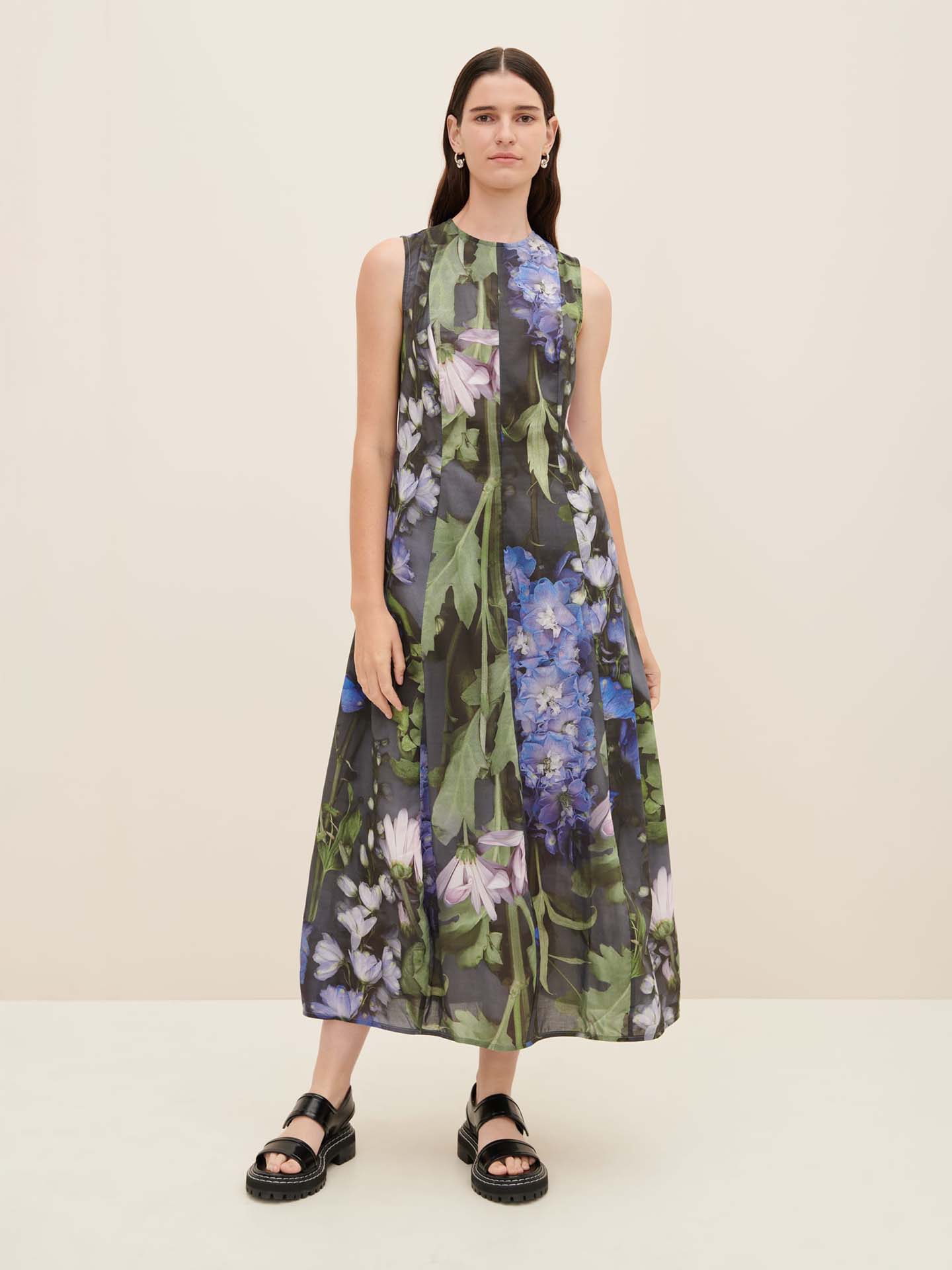 A person wearing the Mosaic Dress by Kowtow, adorned with purple and green floral patterns, stylishly standing on a plain background.