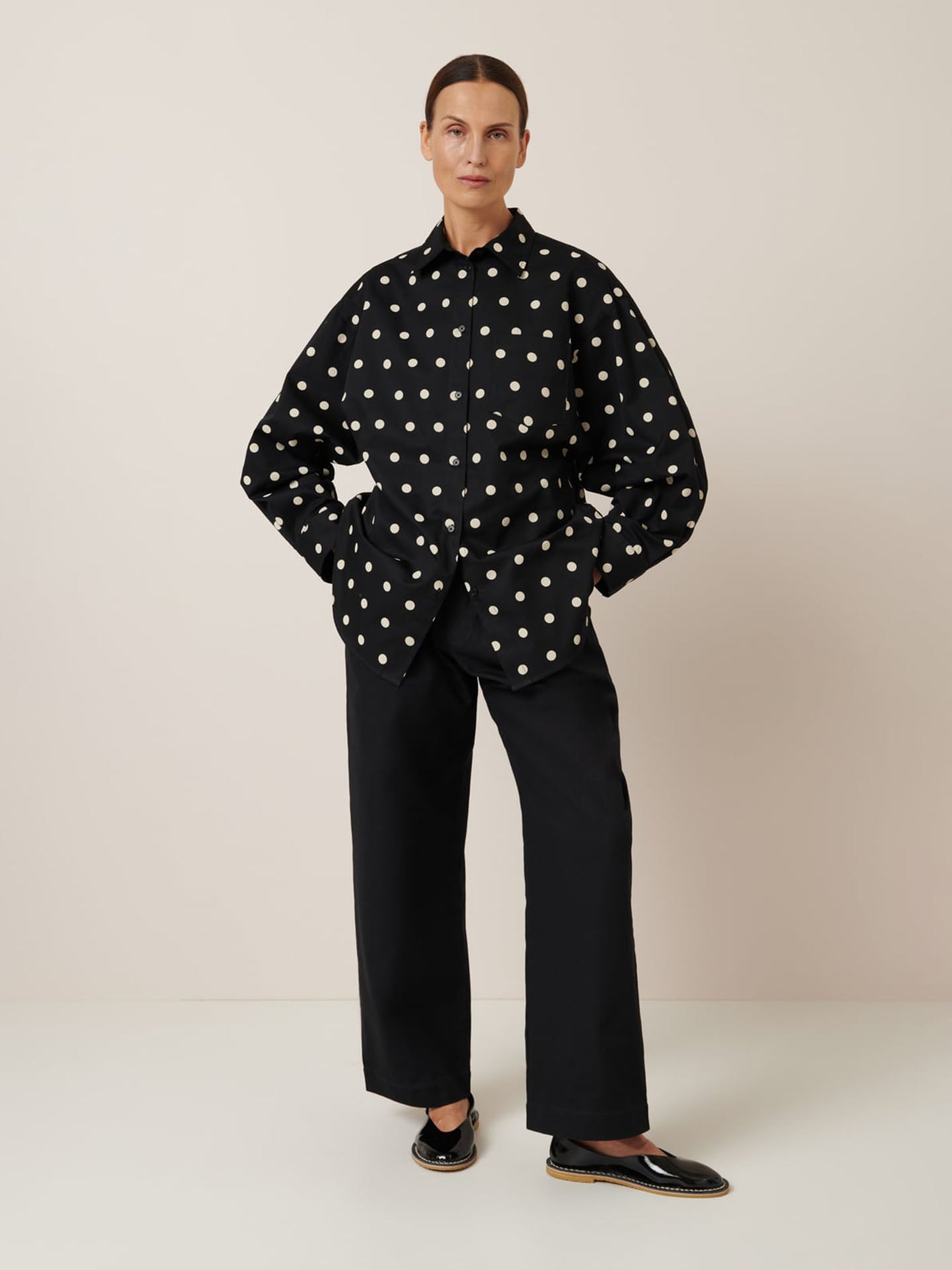 A person stands against a plain background wearing the Kowtow Nova Shirt – Polka Dot, black pants, and black shoes for an effortlessly chic look. For those considering an oversized fit, refer to the size guide to perfect this stylish ensemble.