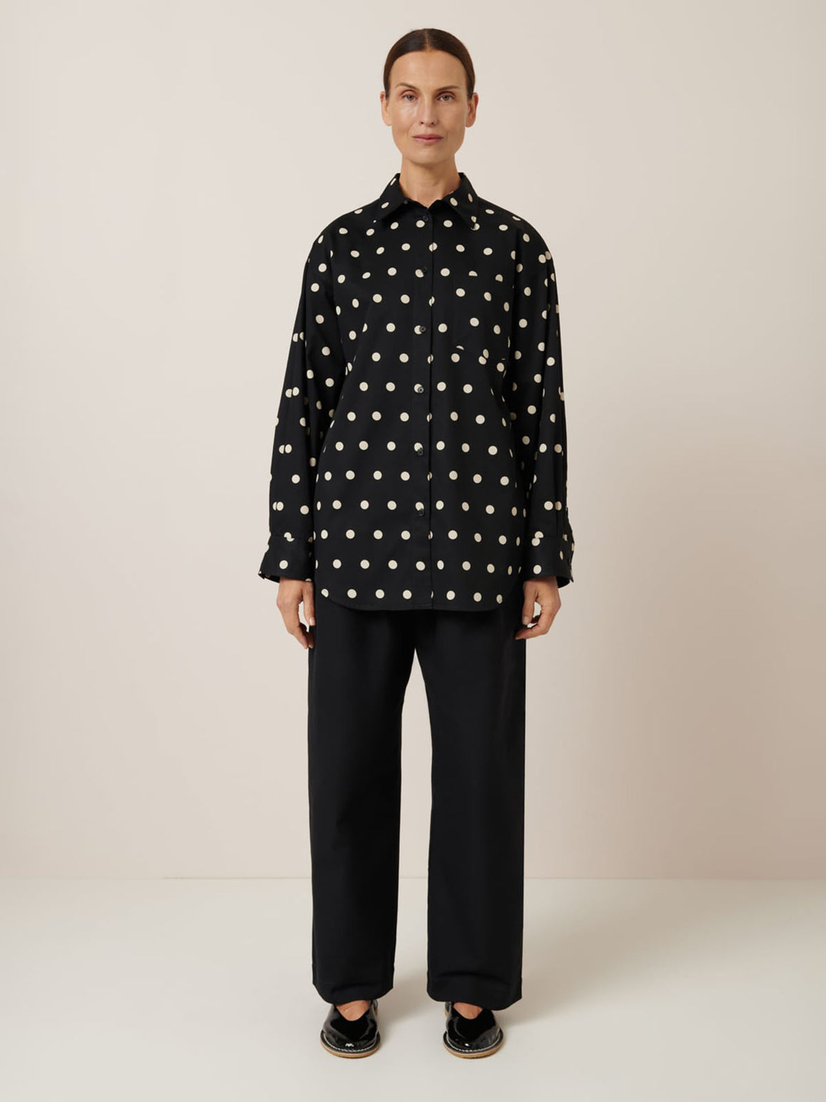 A person stands against a plain background wearing an oversized Nova Shirt – Polka Dot by Kowtow, black pants, and black sandals. With short hair and a neutral expression, they show how playful sizing down can enhance any look.