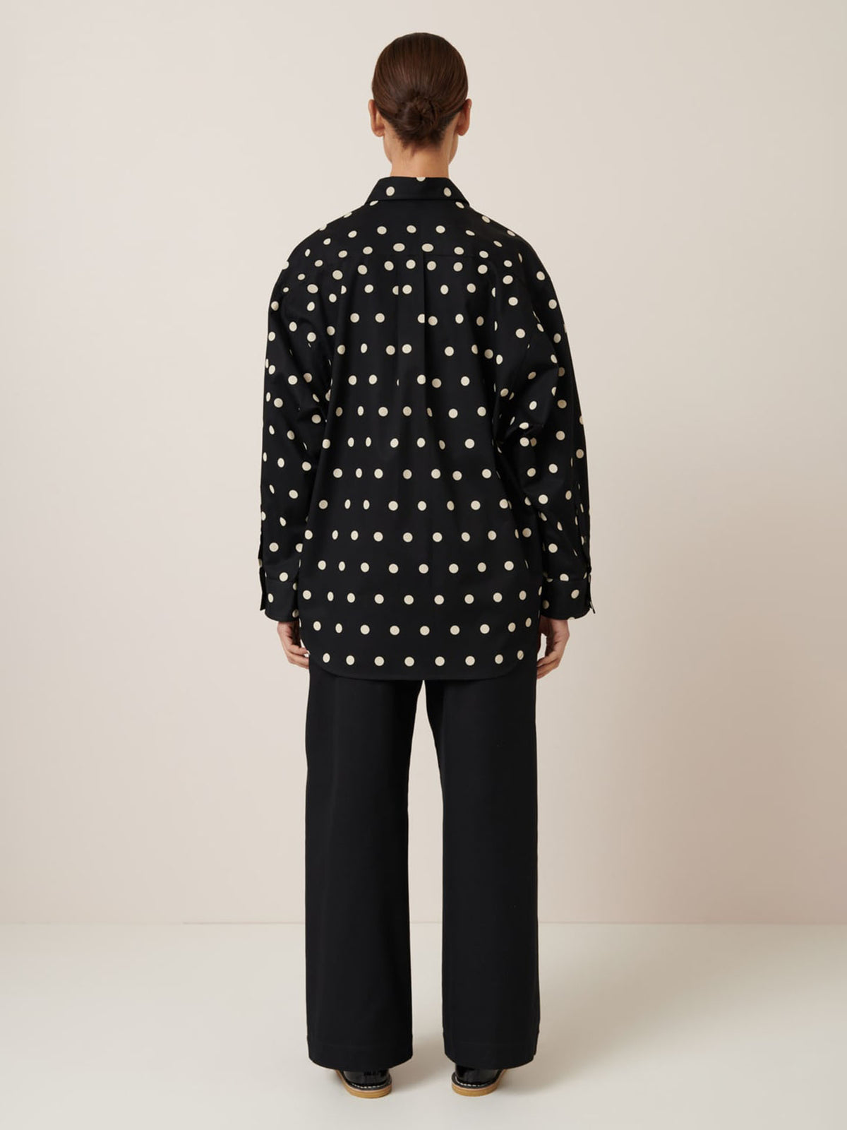 A person in an oversized Nova Shirt – Polka Dot from Kowtow and black pants stands with their back to the camera against a plain background.