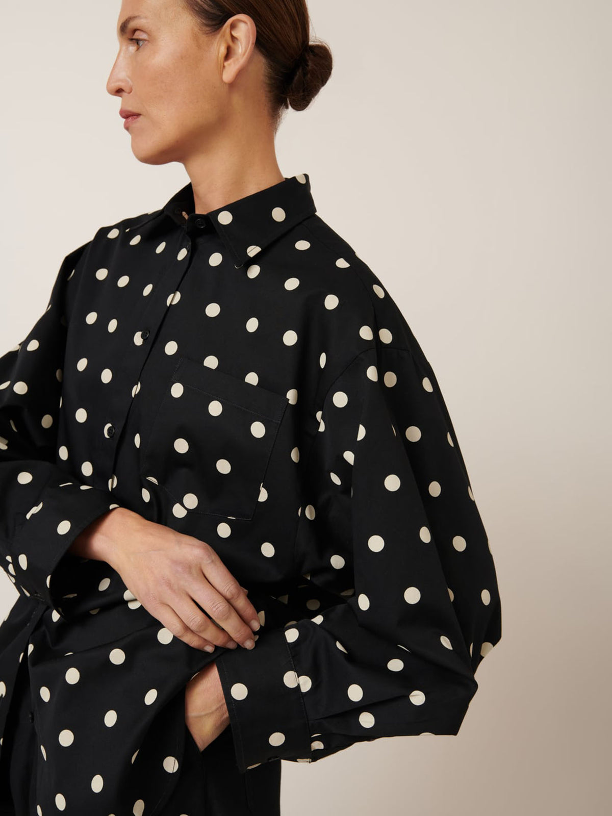 A person in a Nova Shirt – Polka Dot by Kowtow, glancing sideways; their hair is tied back neatly. The oversized shirt adds effortless style.