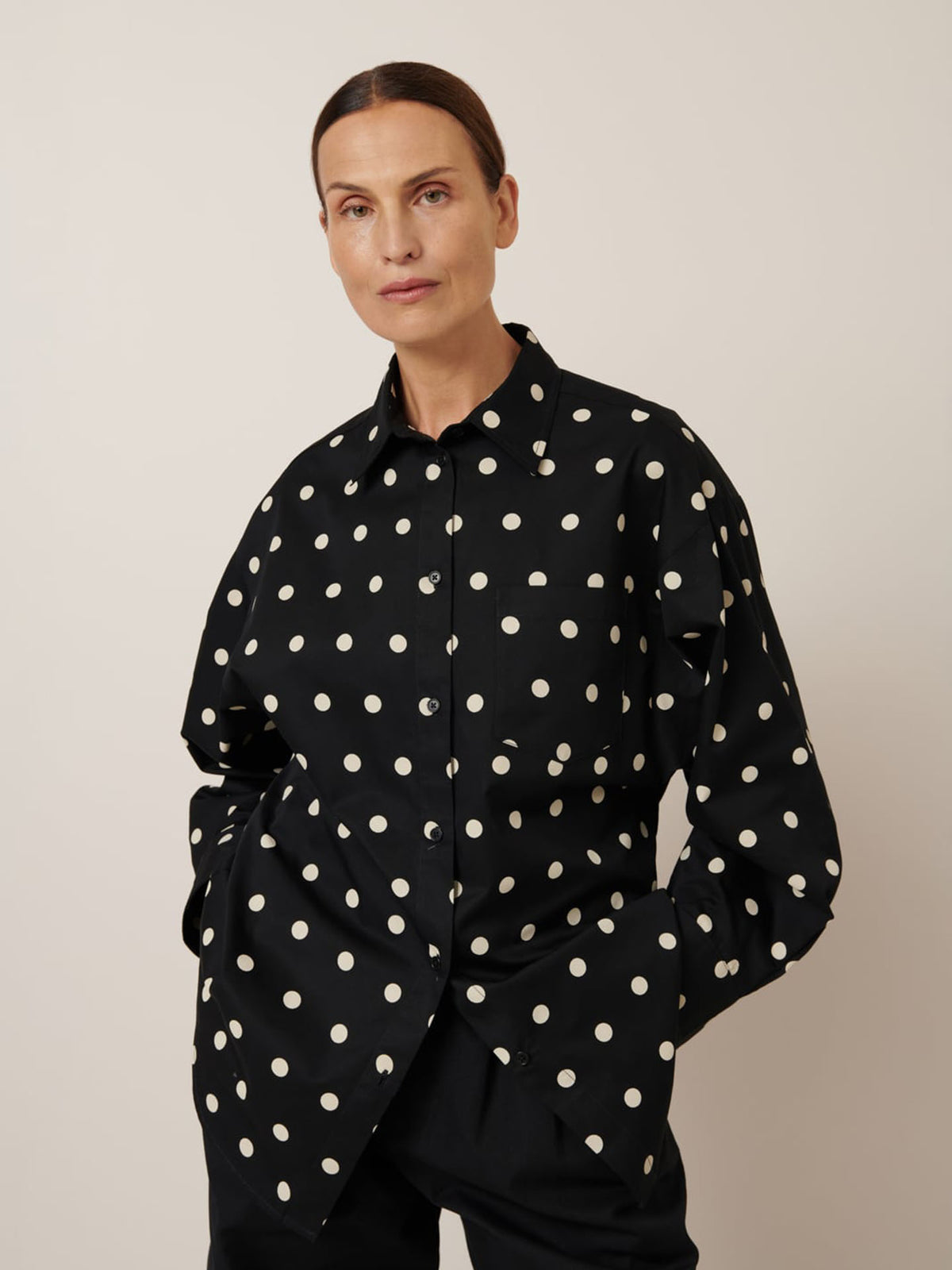 A person models the Kowtow Nova Shirt – an oversized black shirt with white polka dots, hands in pockets against a plain background, demonstrating how opting for this size can still achieve a laid-back yet stylish vibe.