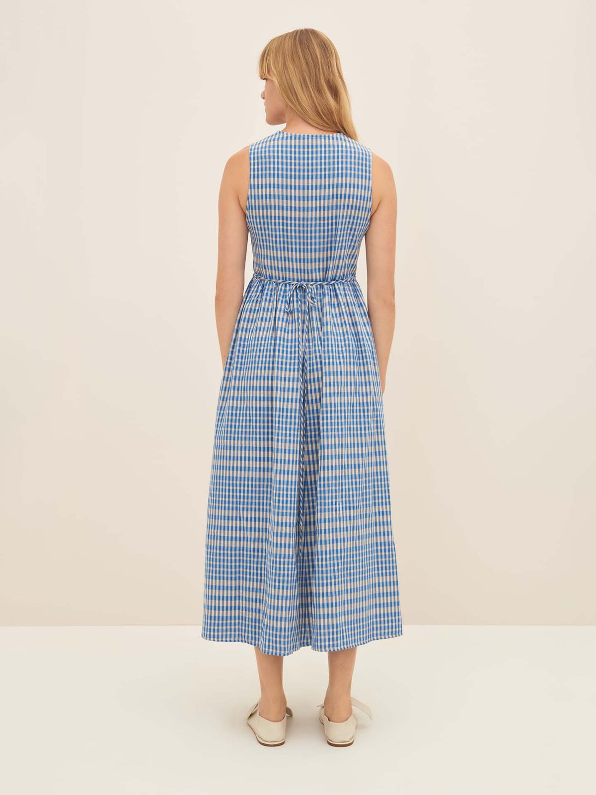 A person wearing the Kowtow Outline Dress, characterized by its blue and white checkered pattern and sleeveless design, stands facing away. The dress, likely in size S, features a gathered waist and is paired with standard fit slip-on shoes.