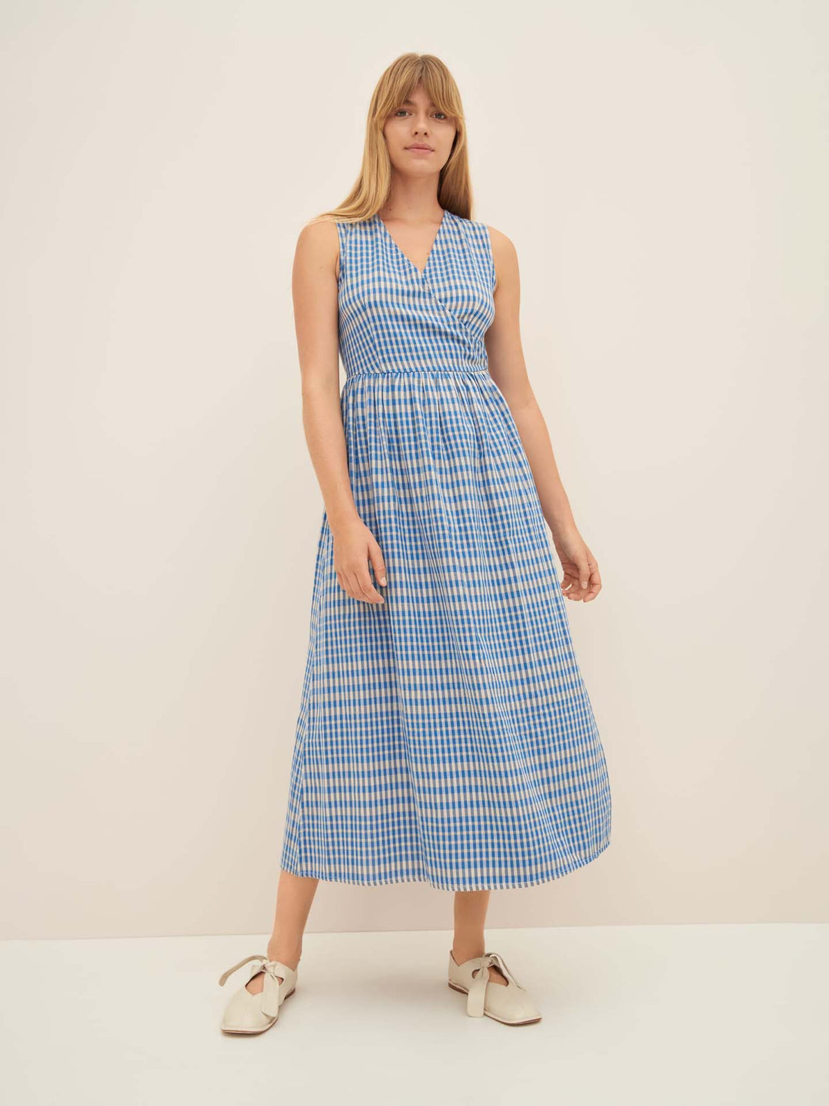 A person stands against a plain background wearing an Outline Dress by Kowtow in blue and white gingham, size S for a standard fit, paired with white slip-on shoes.