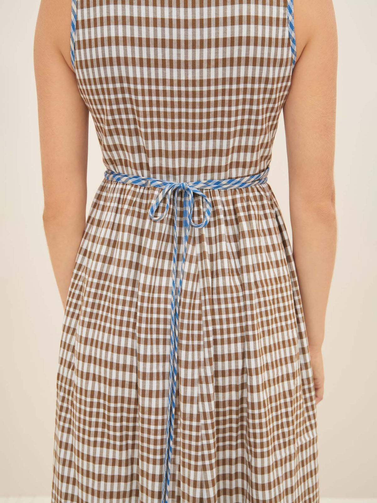 A person wearing the Kowtow Outline Dress in brown and white checkered pattern with blue trim, viewed from the back. The dress in size S is cinched at the waist with a blue tie, ensuring a standard fit.