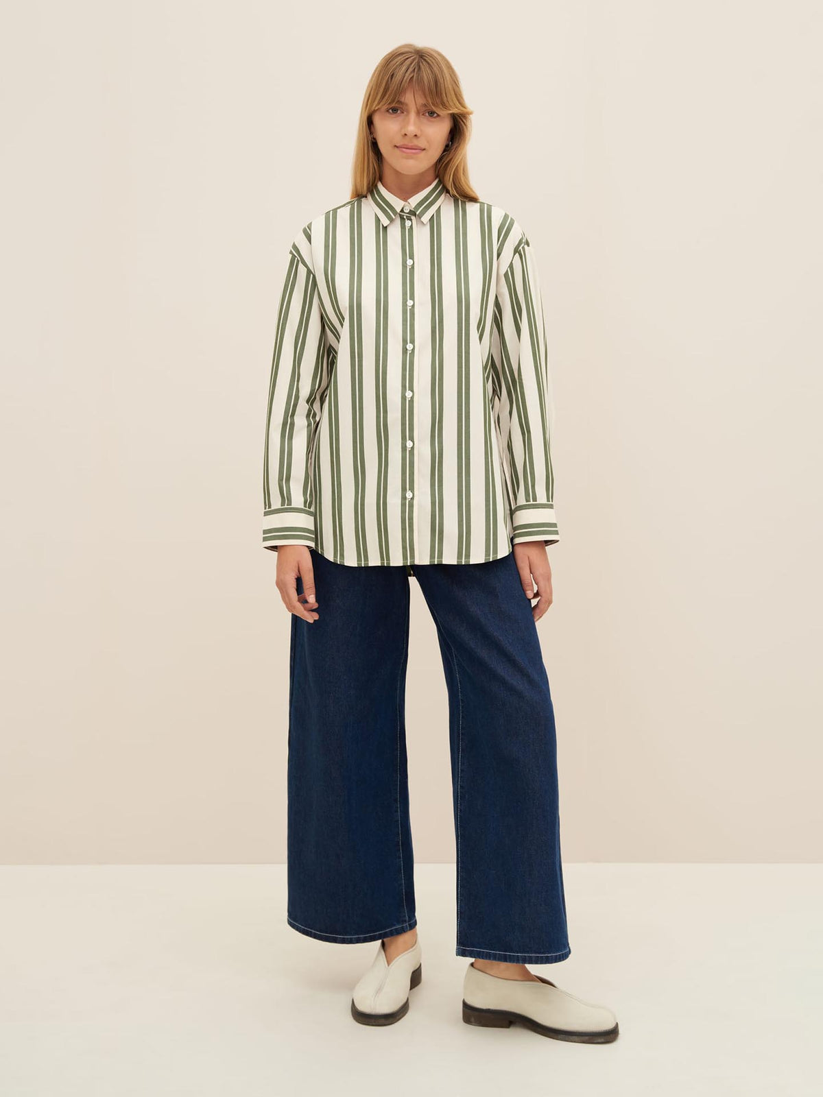 A person is standing against a plain background, wearing the Petra Shirt – Forest Stripe by Kowtow, which features white and green vertical stripes with an oversized fit, paired with blue wide-leg jeans and white shoes.