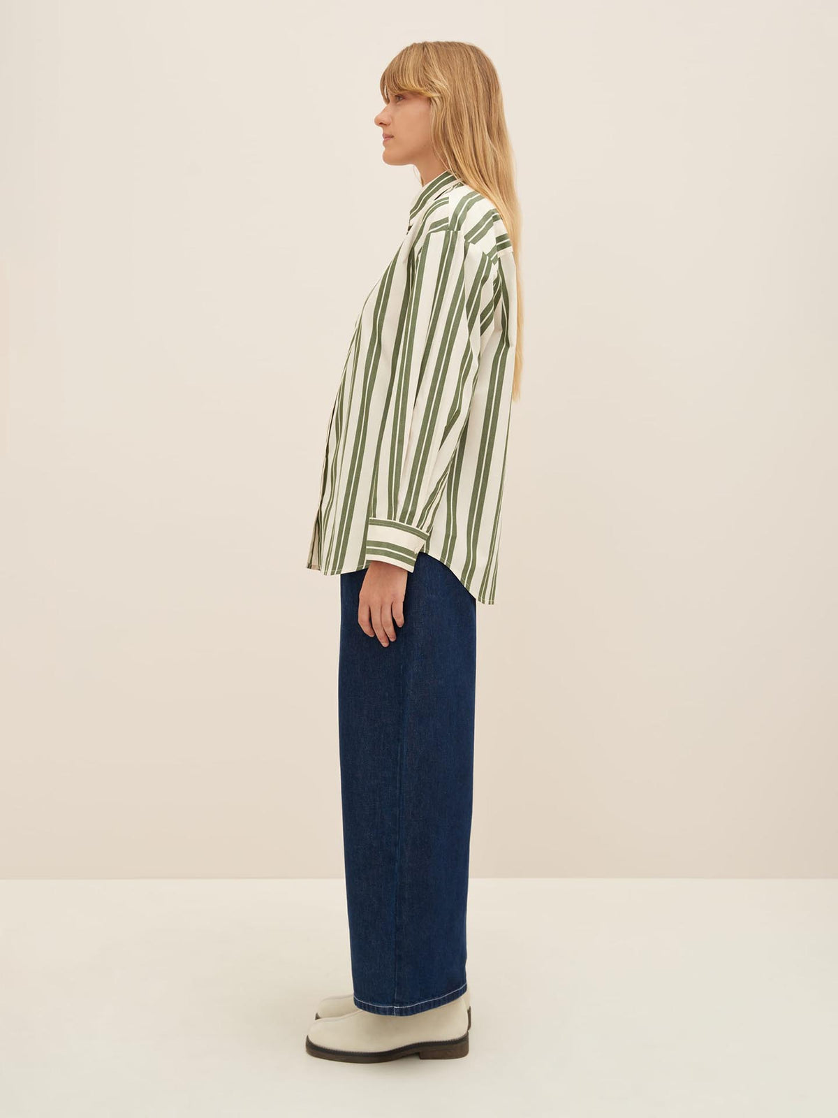 A person with long hair is standing in profile, showcasing an oversized fit with the Petra Shirt – Forest Stripe by Kowtow, blue wide-leg pants, and white shoes against a plain background.