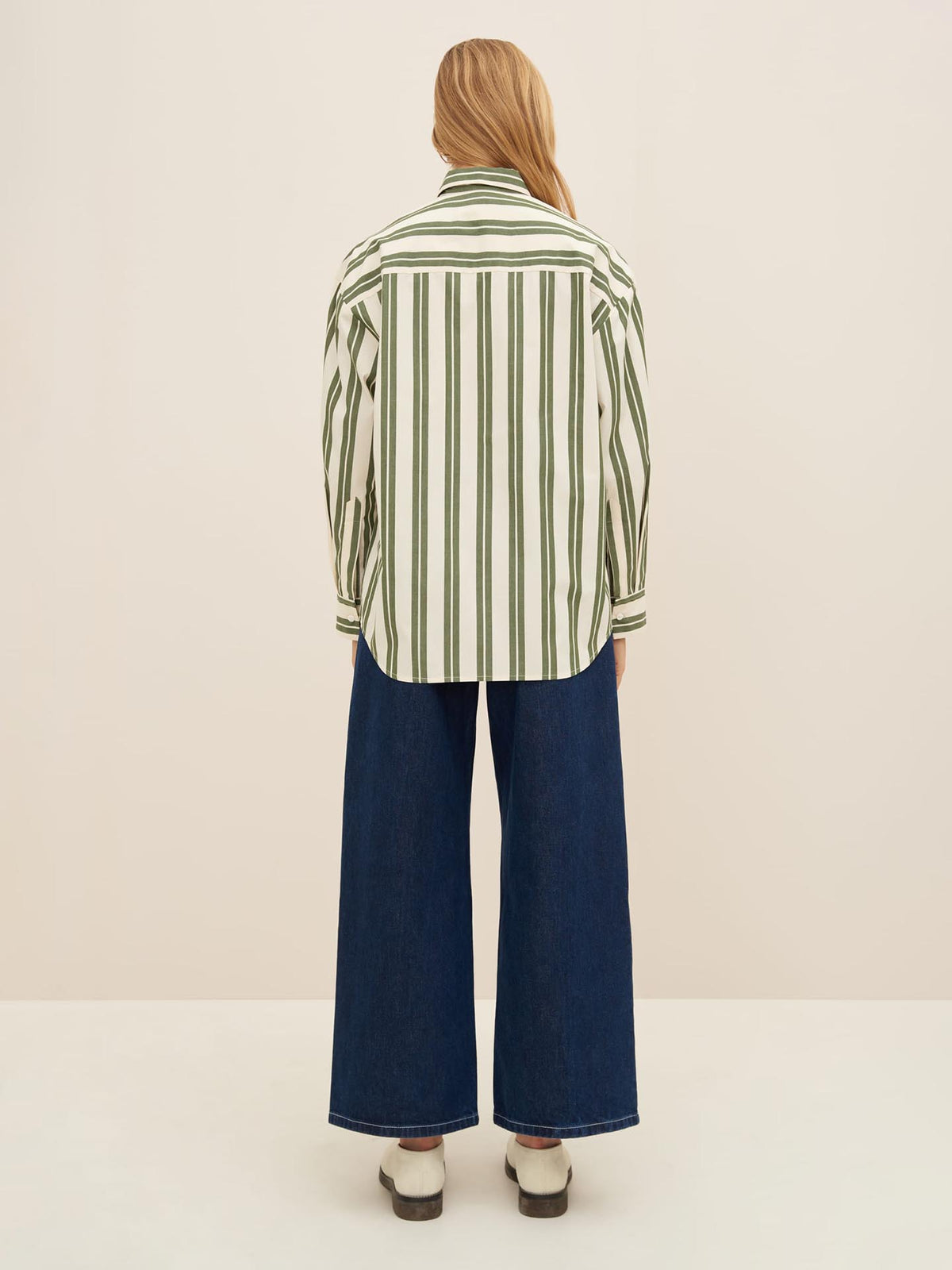 A person with long hair is standing with their back to the camera, wearing an oversized Kowtow Petra Shirt in Forest Stripe, wide-leg blue jeans, and white shoes.