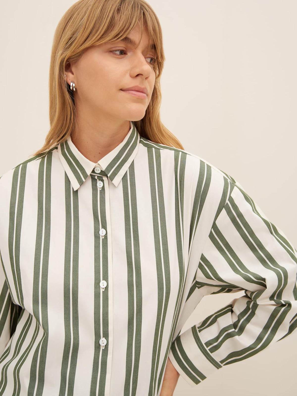 A person with long hair wears the Petra Shirt – Forest Stripe, an oversized fit green and white striped button-up shirt from Kowtow, and looks to the side.