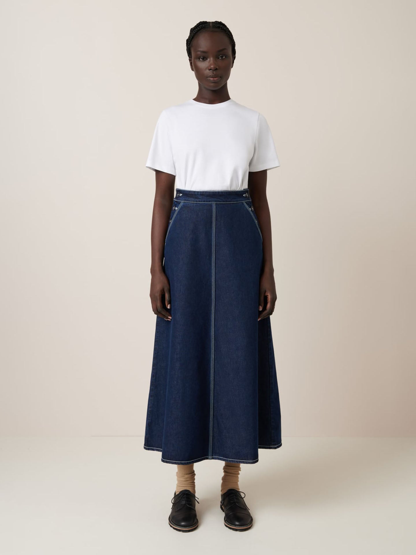 A person is pictured in front of a plain backdrop wearing Kowtow's Piper Skirt in Indigo Denim, paired with a relaxed-fit white t-shirt, beige socks, and black shoes.