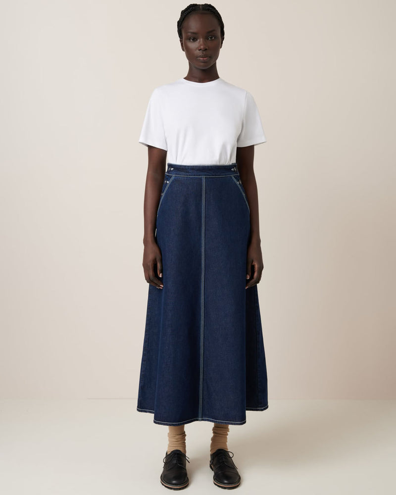 A person is pictured in front of a plain backdrop wearing Kowtow's Piper Skirt in Indigo Denim, paired with a relaxed-fit white t-shirt, beige socks, and black shoes.