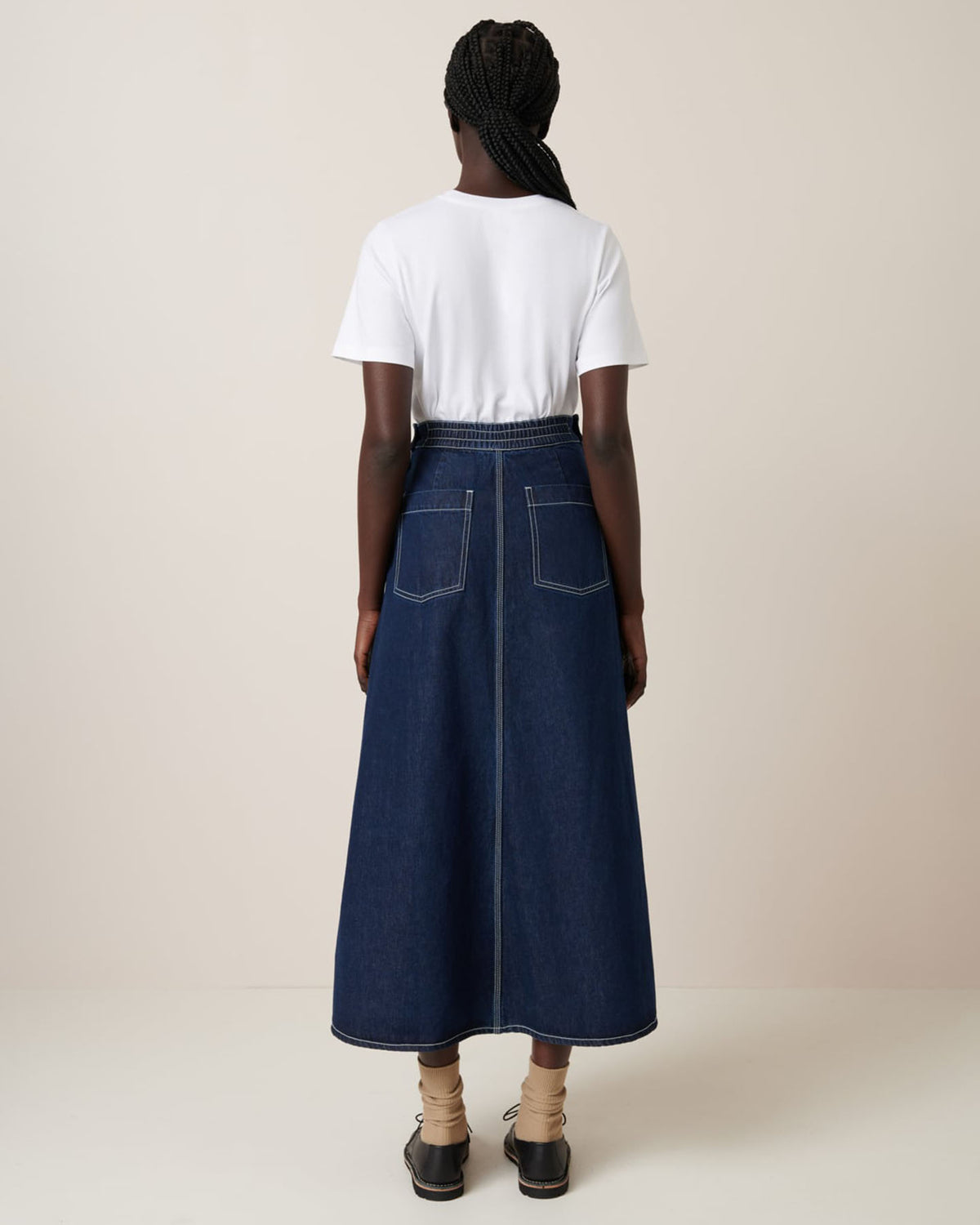 A person with braided hair is wearing a white t-shirt and the Piper Skirt – Indigo Denim by Kowtow, showcasing a relaxed fit from the back.