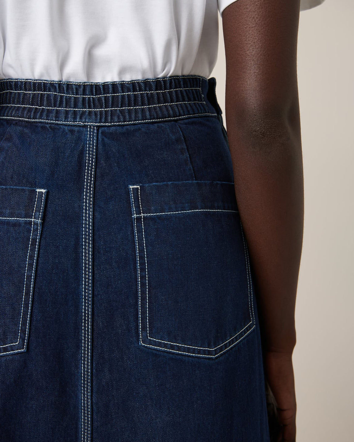 A person is seen from behind wearing a Kowtow Piper Skirt – Indigo Denim, featuring a relaxed fit with back pockets, paired with a white shirt.