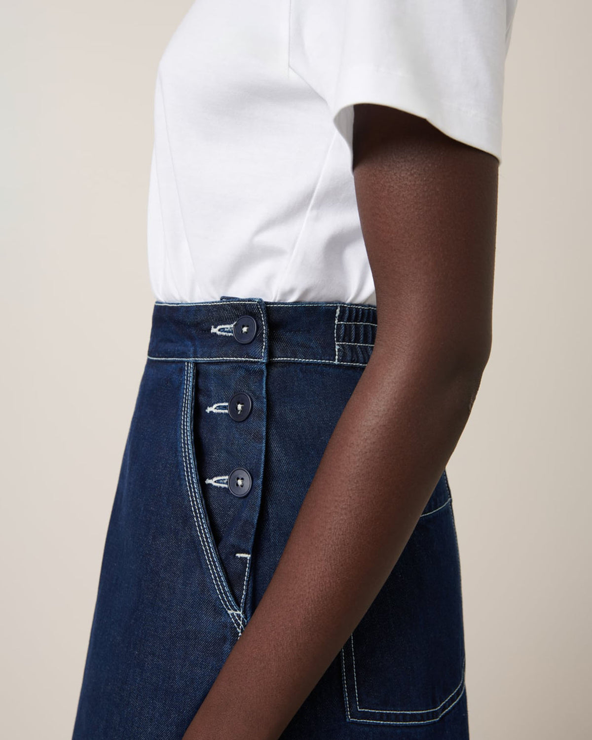 A person is wearing a white shirt and the Kowtow Piper Skirt in Indigo Denim, featuring side button details and offering a relaxed fit.