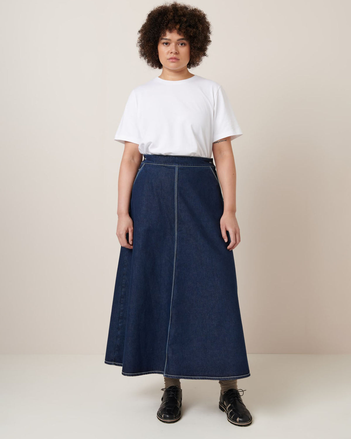 Dressed in a relaxed fit white T-shirt paired with Kowtow's "Piper Skirt – Indigo Denim," a person stands against a plain background, effortlessly showcasing comfort and style.