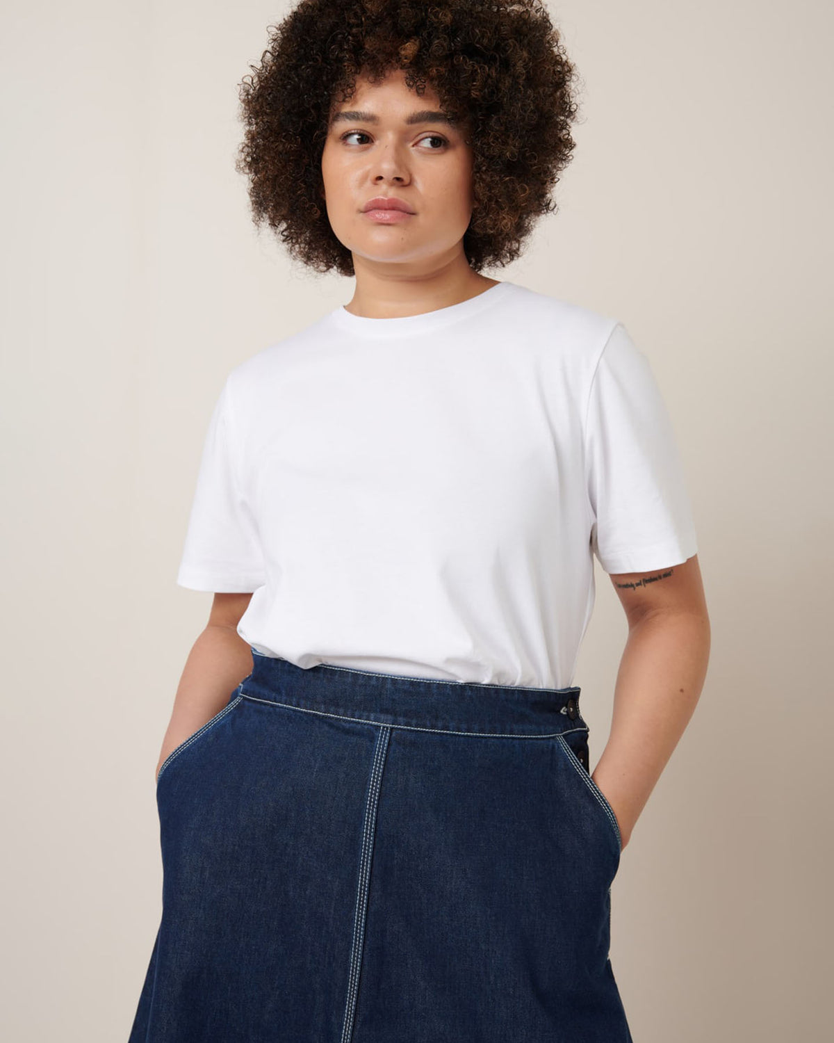 A person with curly hair wearing a Kowtow relaxed fit white T-shirt and the Piper Skirt in Indigo Denim stands with hands in pockets against a neutral backdrop.