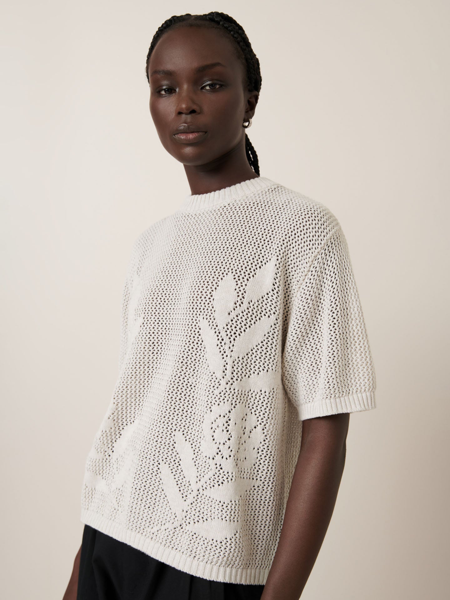 A person stands against a plain background, modeling the Kowtow Rosa Tee in Oat Melange, a short-sleeved knit sweater with a charming floral pattern.