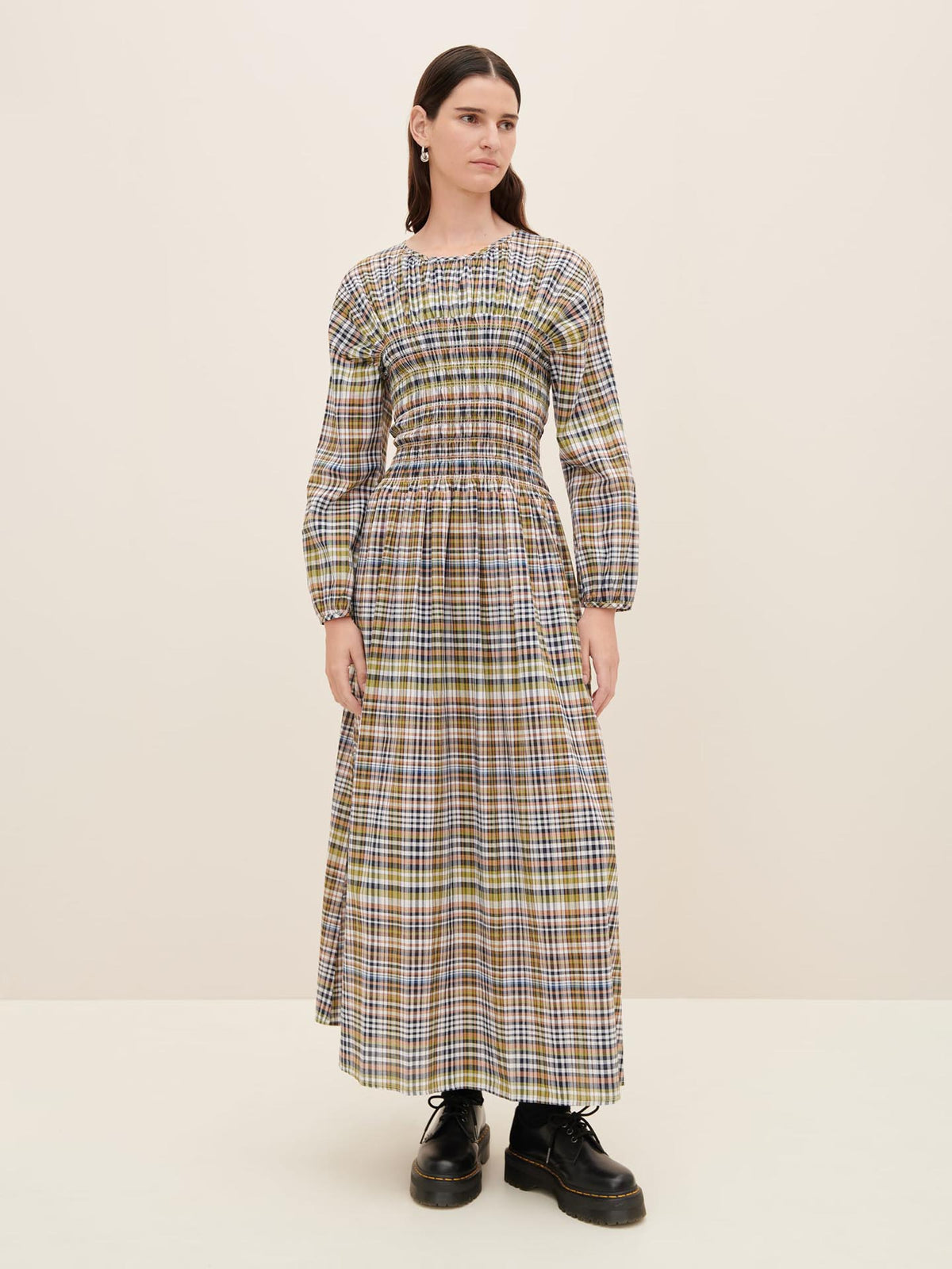 A person stands against a plain background wearing the stylish Sienna Dress by Kowtow, featuring a long plaid design with a shirred waist and long sleeves, paired with black platform shoes to highlight its standard fit. For more details, refer to the size guide and product measurements.