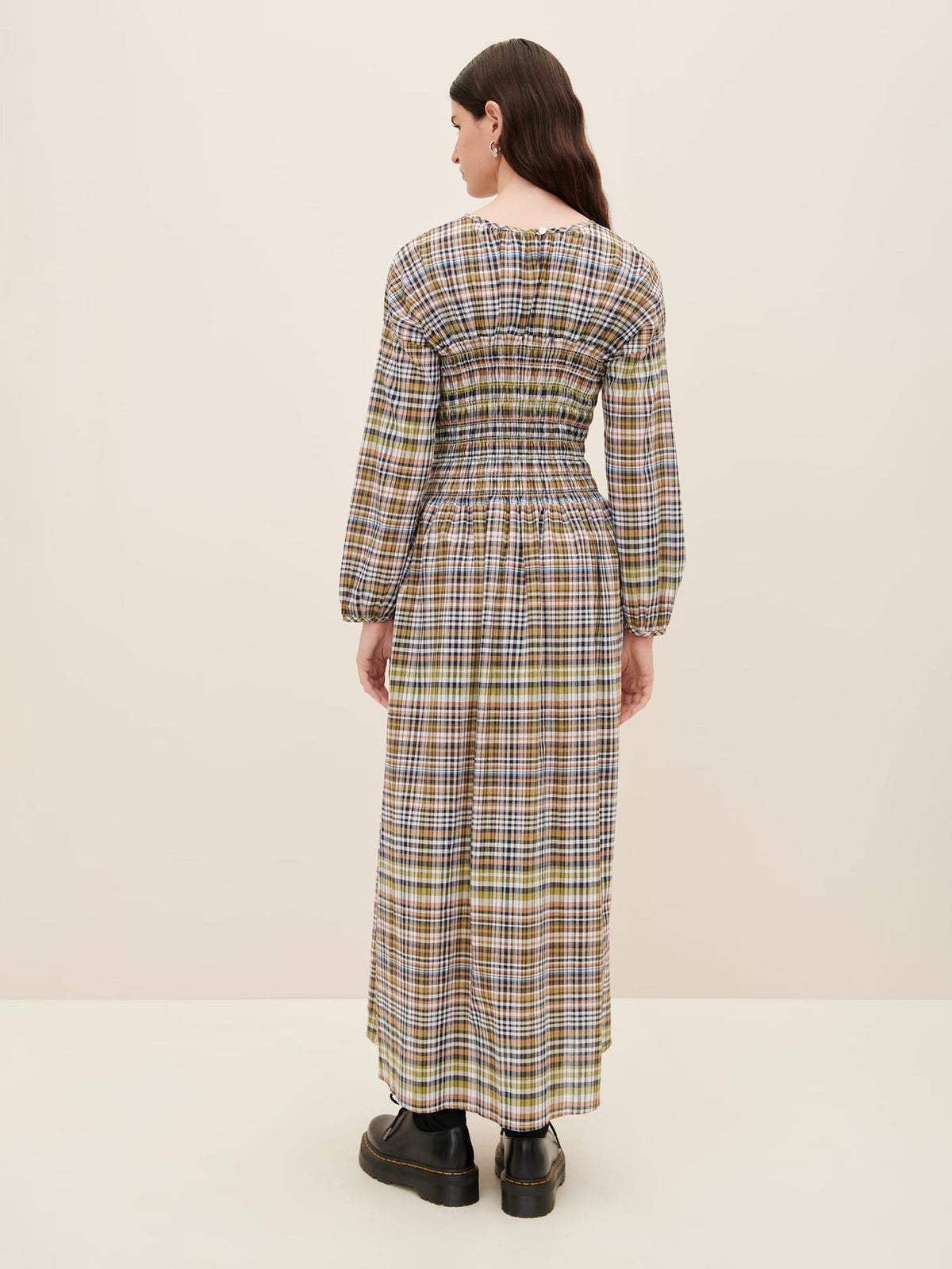 Person wearing the Kowtow Sienna Dress, a long, multi-color plaid garment with long sleeves, seen from the back. The standard fit accentuates their figure, complemented by black platform shoes.