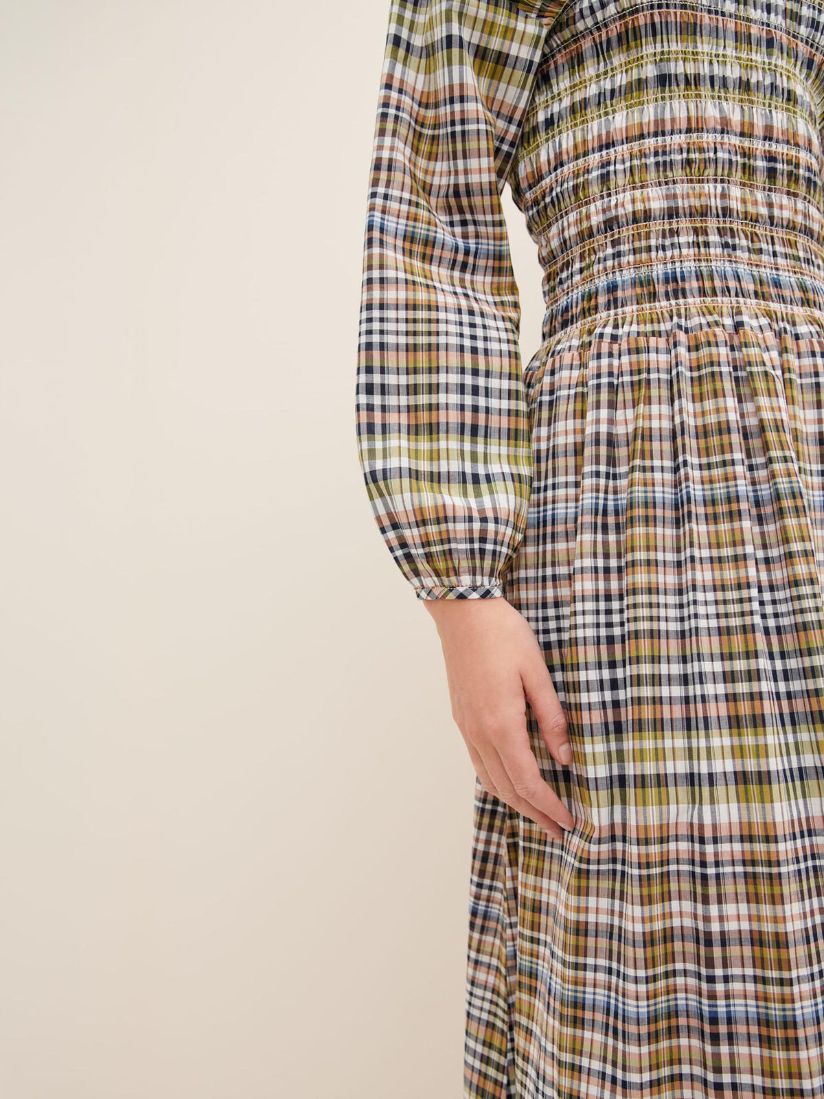 Close-up of a person&#39;s hand resting by their side, wearing the Kowtow Sienna Dress, a plaid, long-sleeved design with smocking at the waist and sleeves, set against a plain background. For the perfect Standard Fit, consult our Size Guide.