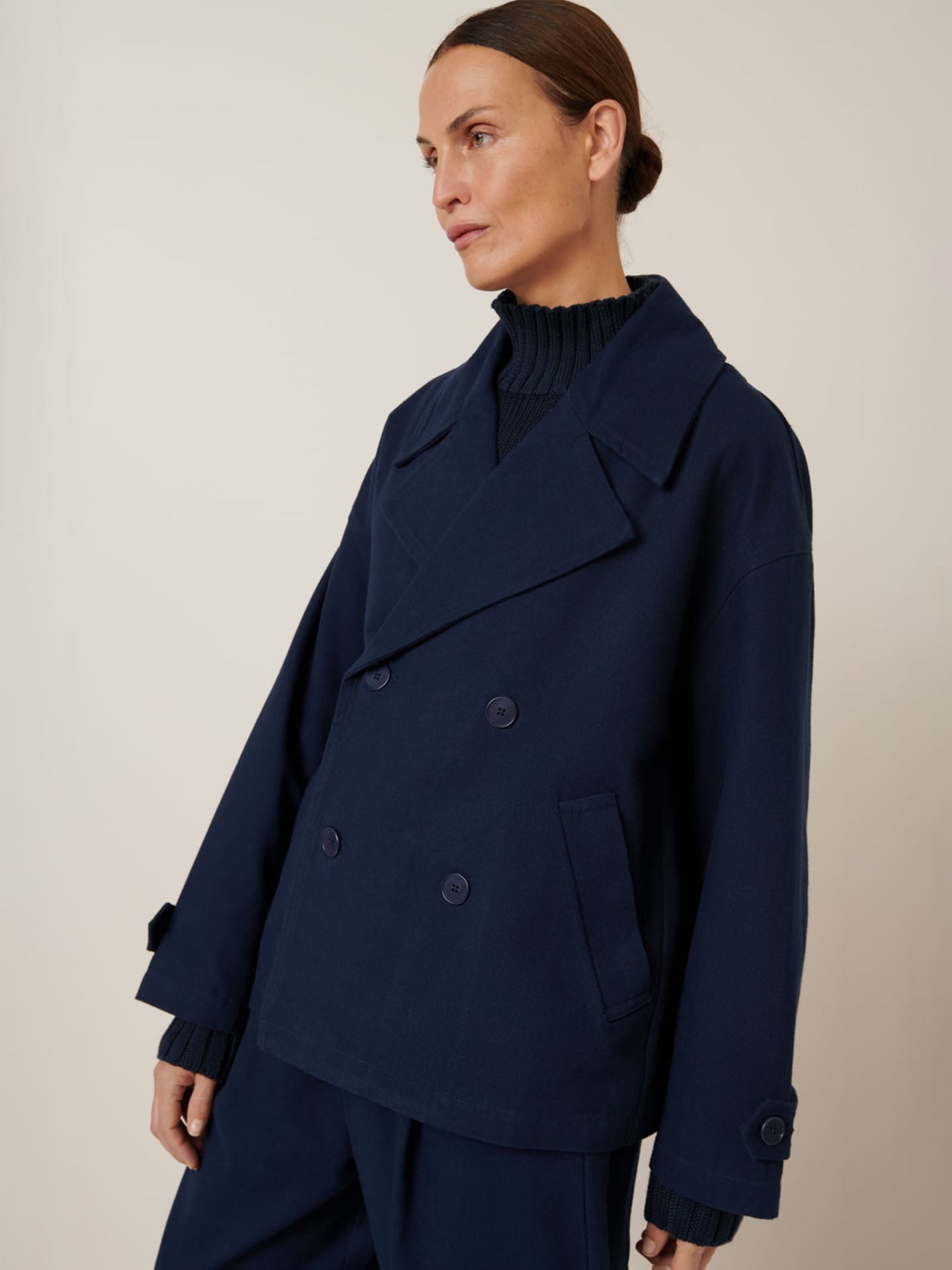 A person wearing Kowtow's Silhouette Jacket in Evening Navy, featuring a double-breasted design with an oversized fit and paired with a high-neck sweater, stands against a plain background, looking to the side.