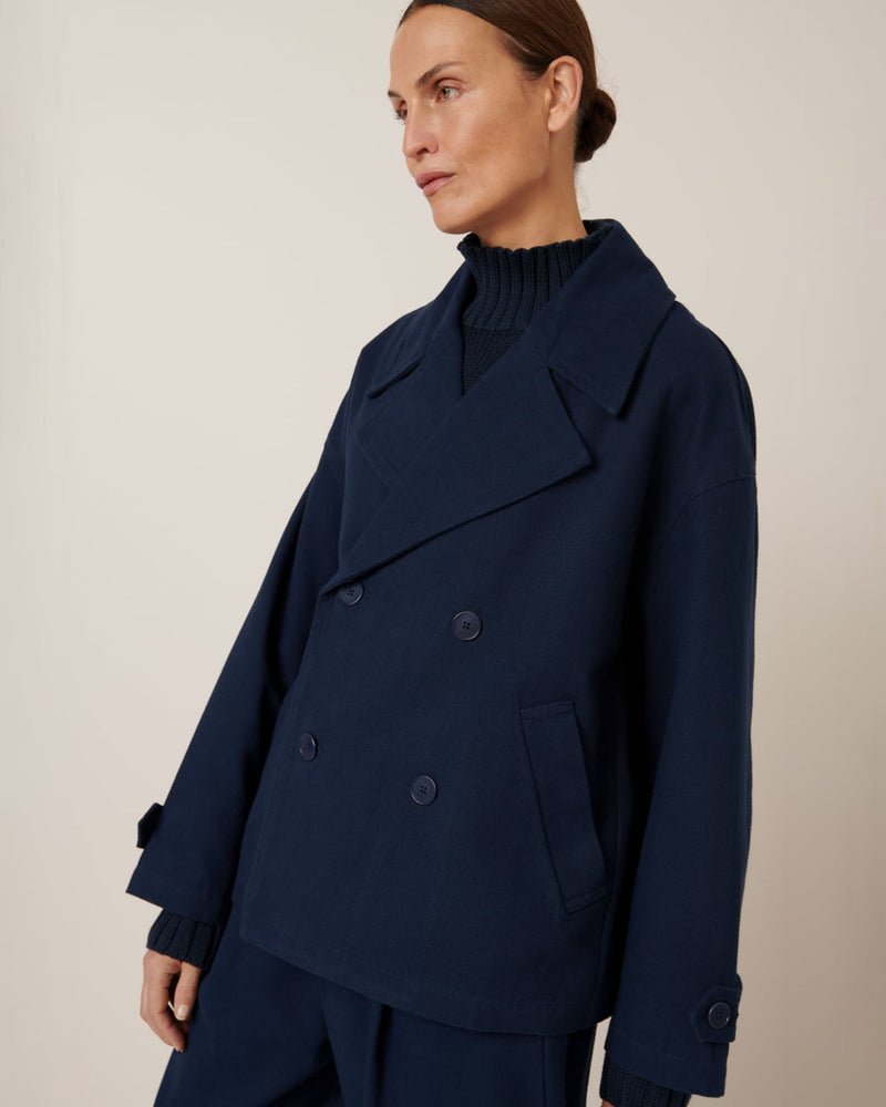 A person wearing Kowtow's Silhouette Jacket in Evening Navy, featuring a double-breasted design with an oversized fit and paired with a high-neck sweater, stands against a plain background, looking to the side.