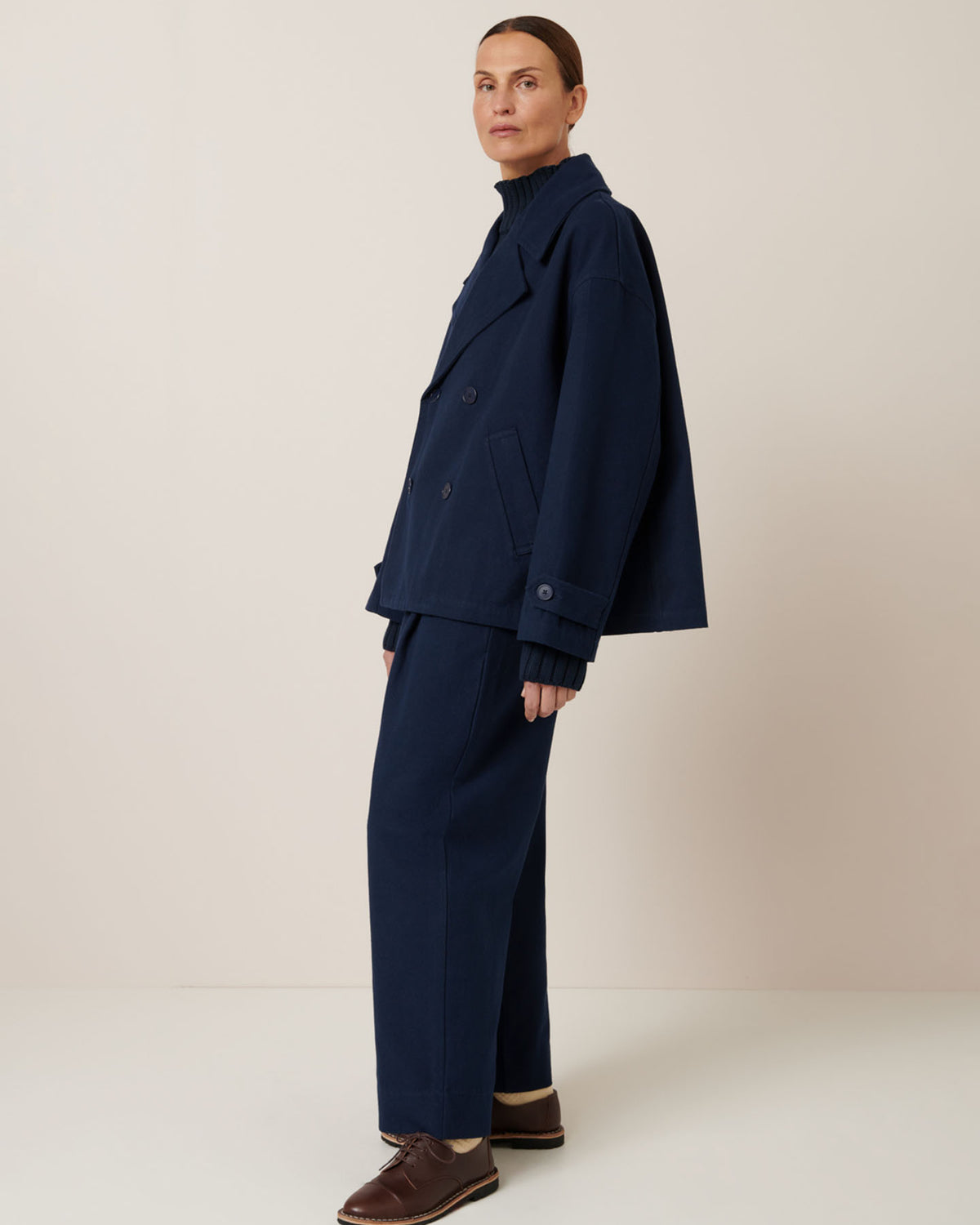 A person models an oversized fit with the Kowtow Silhouette Jacket in Evening Navy paired with matching pants and brown shoes, all set against a plain beige background.