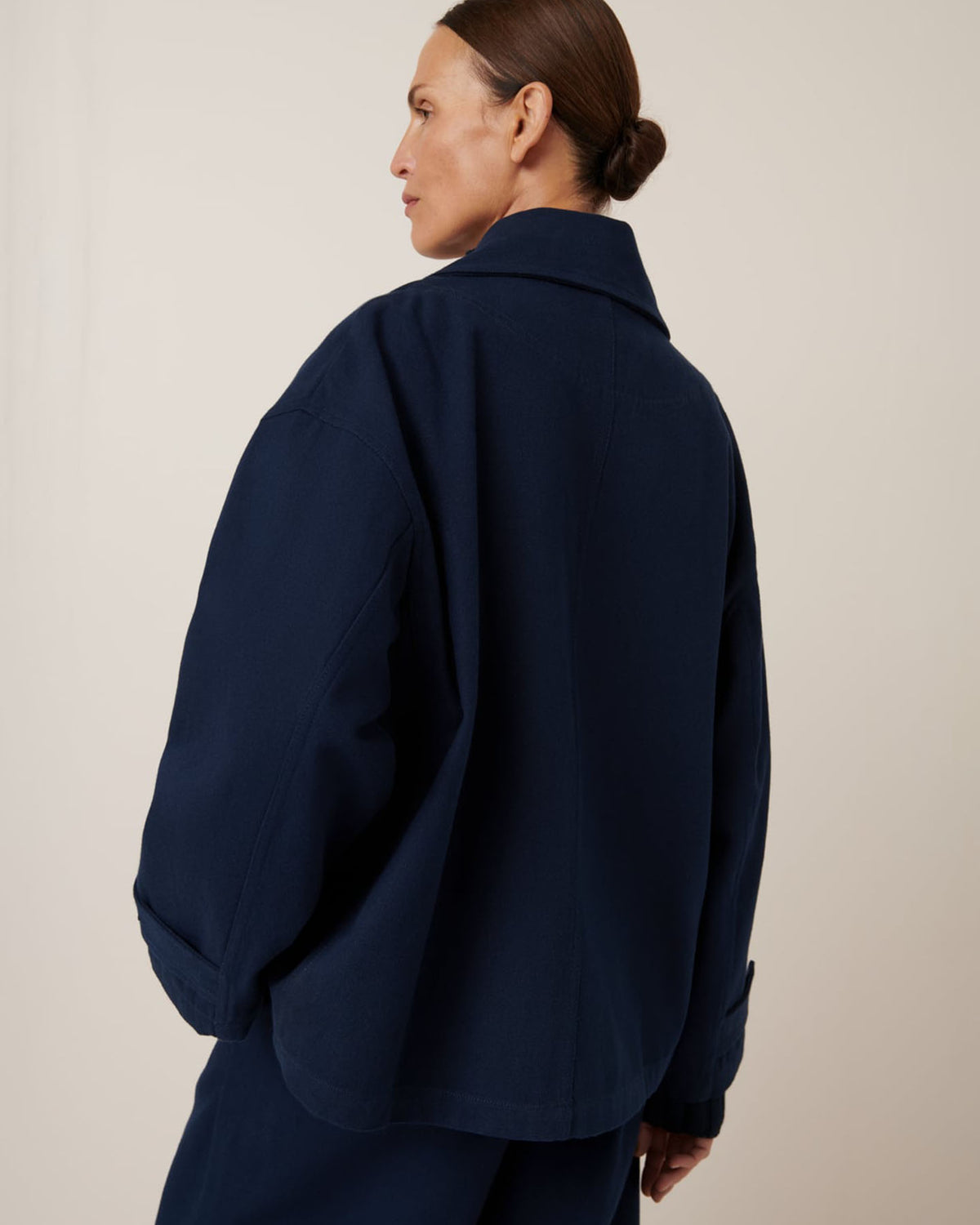 A person stands slightly turned away, highlighting the oversized fit of Kowtow's Silhouette Jacket in Evening Navy. The garment pattern adds subtle elegance against the simple, neutral backdrop.