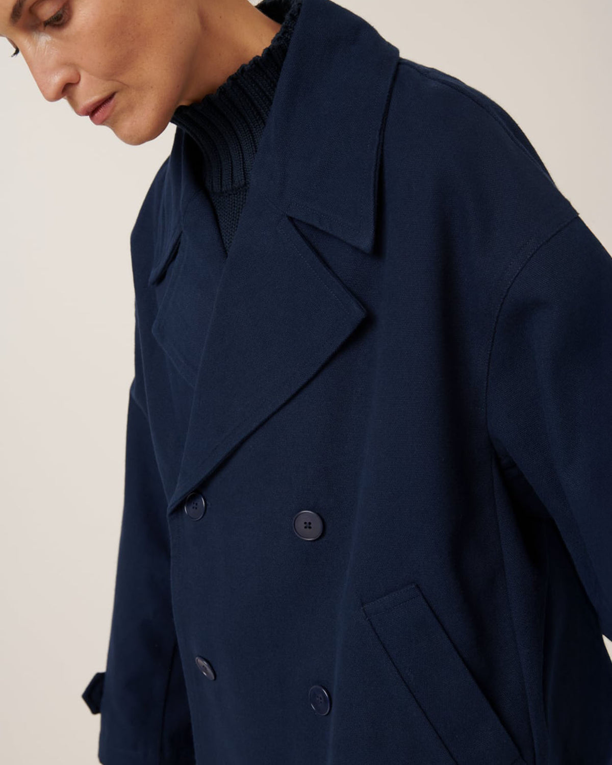 A person draped in the oversized Silhouette Jacket – Evening Navy by Kowtow over a black turtleneck sweater glances downward against a light background.