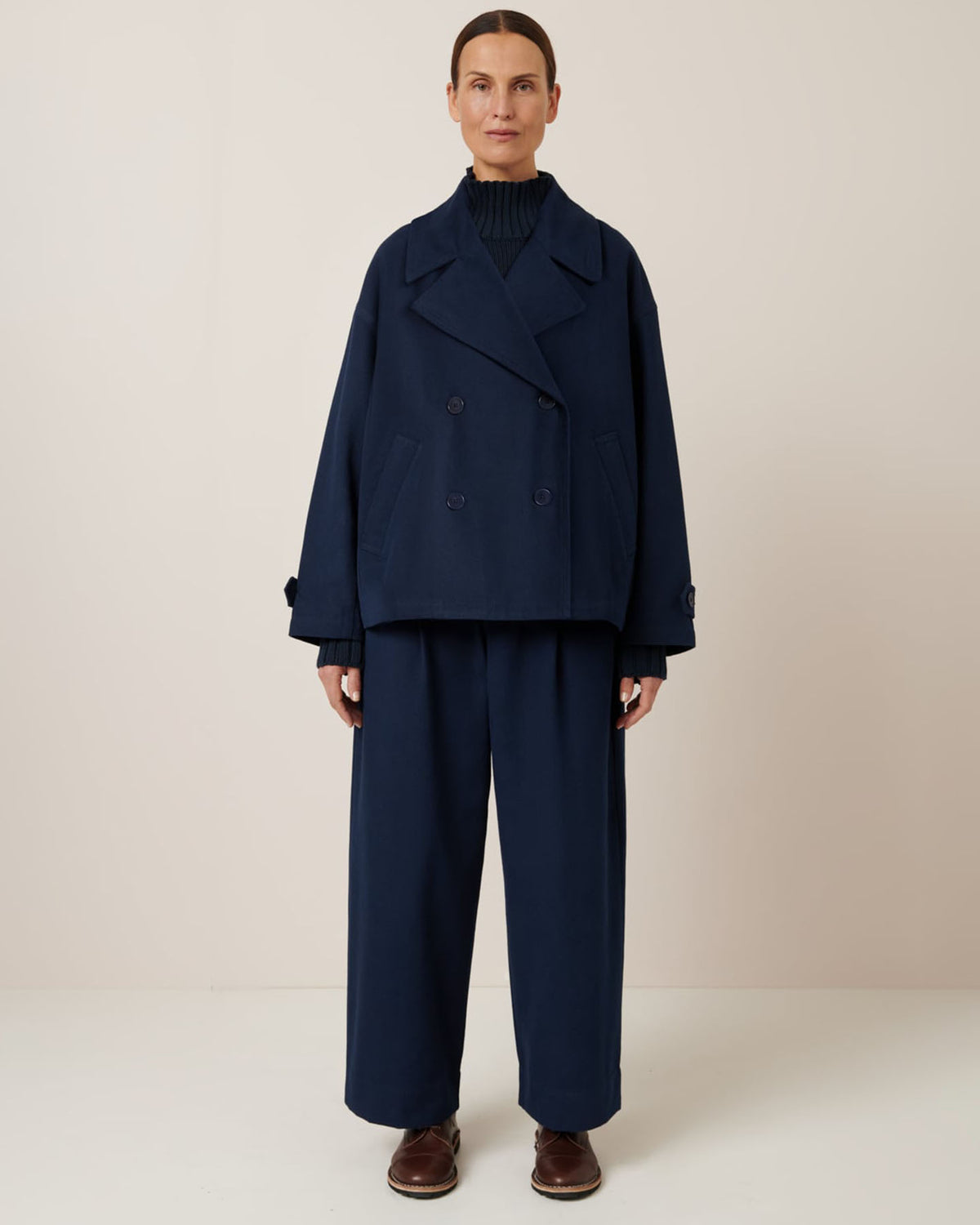 A person stands against a neutral background wearing the Silhouette Jacket – Evening Navy by Kowtow, with matching wide-leg trousers, a turtleneck, and dark shoes, showcasing the garment's elegant pattern.
