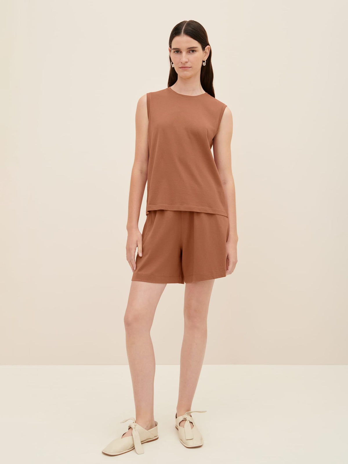 A woman is standing against a plain background wearing the Kowtow Singlet Top in Sienna, matching sienna shorts, and beige slip-on shoes, offering a relaxed fit that&#39;s perfect for any casual occasion. Check the size guide to find your true size.