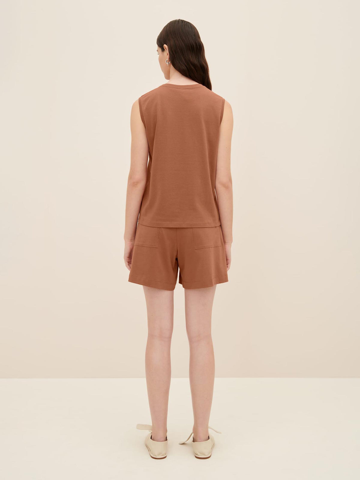 A person with long hair is standing and facing away, wearing the Singlet Top – Sienna by Kowtow paired with matching relaxed-fit shorts and beige slip-on shoes against a neutral background.