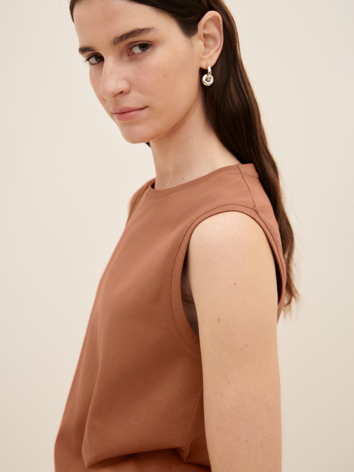 A woman with long dark hair is wearing the sleeveless Singlet Top – Sienna with a relaxed fit by Kowtow and earrings. She is standing against a plain, light-colored background.