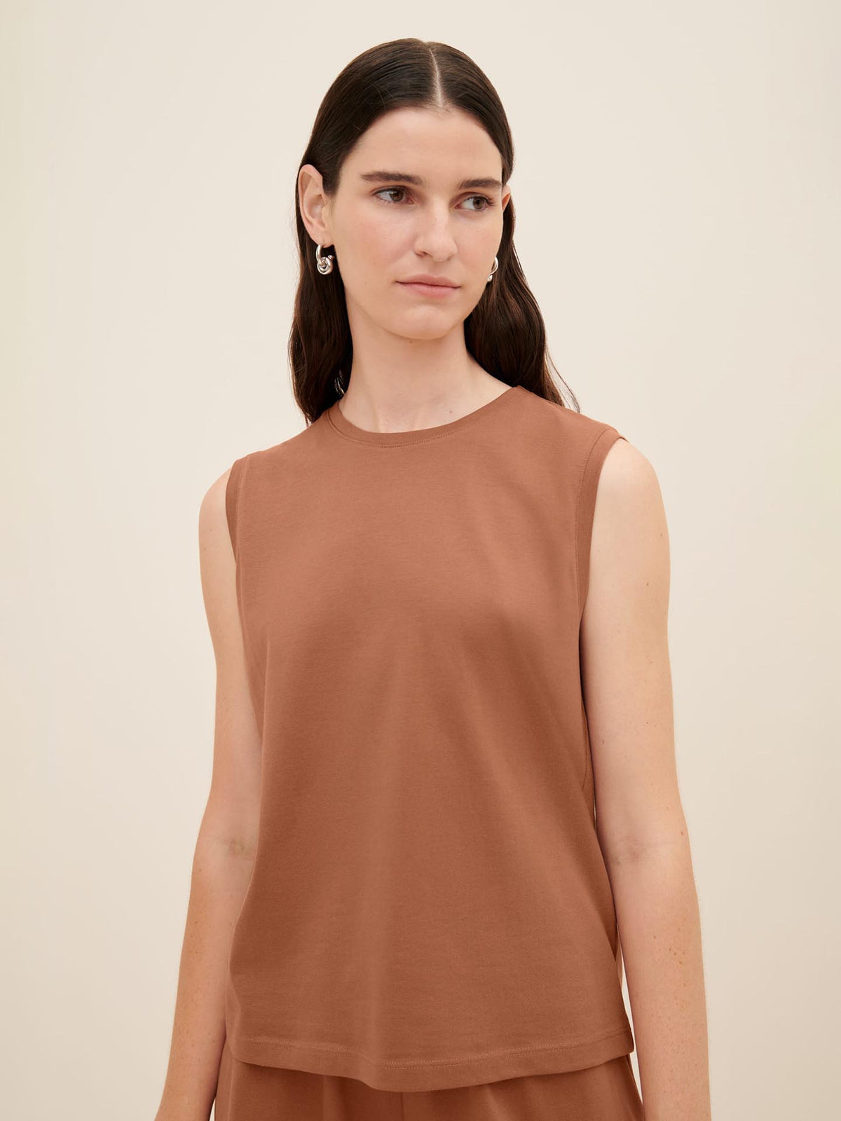 A woman models the Singlet Top – Sienna by Kowtow. The sleeveless top features a relaxed fit and a warm sienna hue. She gazes slightly to the right, her long, straight hair cascading freely, and is captured from the waist up against a plain background.