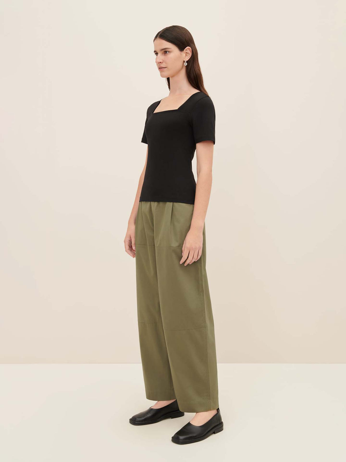 A woman dressed in a black Square Neck Tee by Kowtow and green oversized wide-leg pants stands against a plain light-colored background.