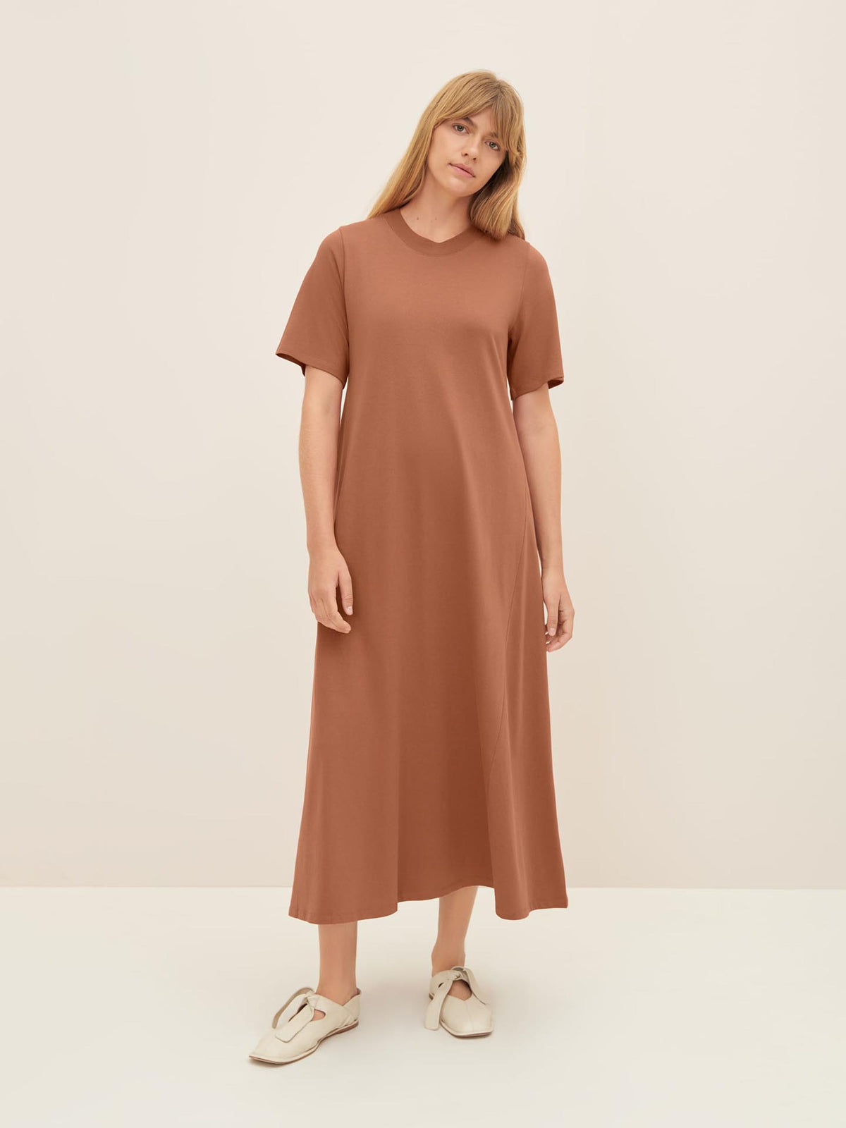 A woman stands against a plain background wearing the Kowtow T-Shirt Swing Dress – Sienna paired with white sneakers. For a comfortable look, refer to the standard fit and consult the Kowtow size guide for detailed product measurements.