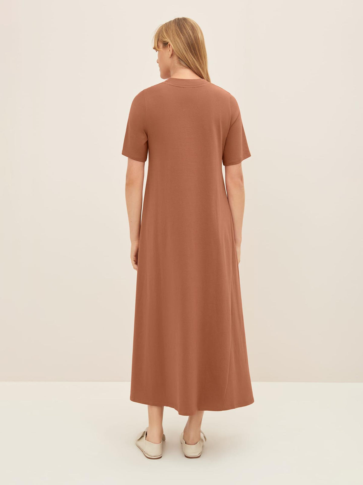 A person with long hair stands facing away, wearing the Kowtow T-Shirt Swing Dress in Sienna matched with white shoes, set against a plain light background. For accurate sizing, please consult the Kowtow size guide for standard fit and product measurements.