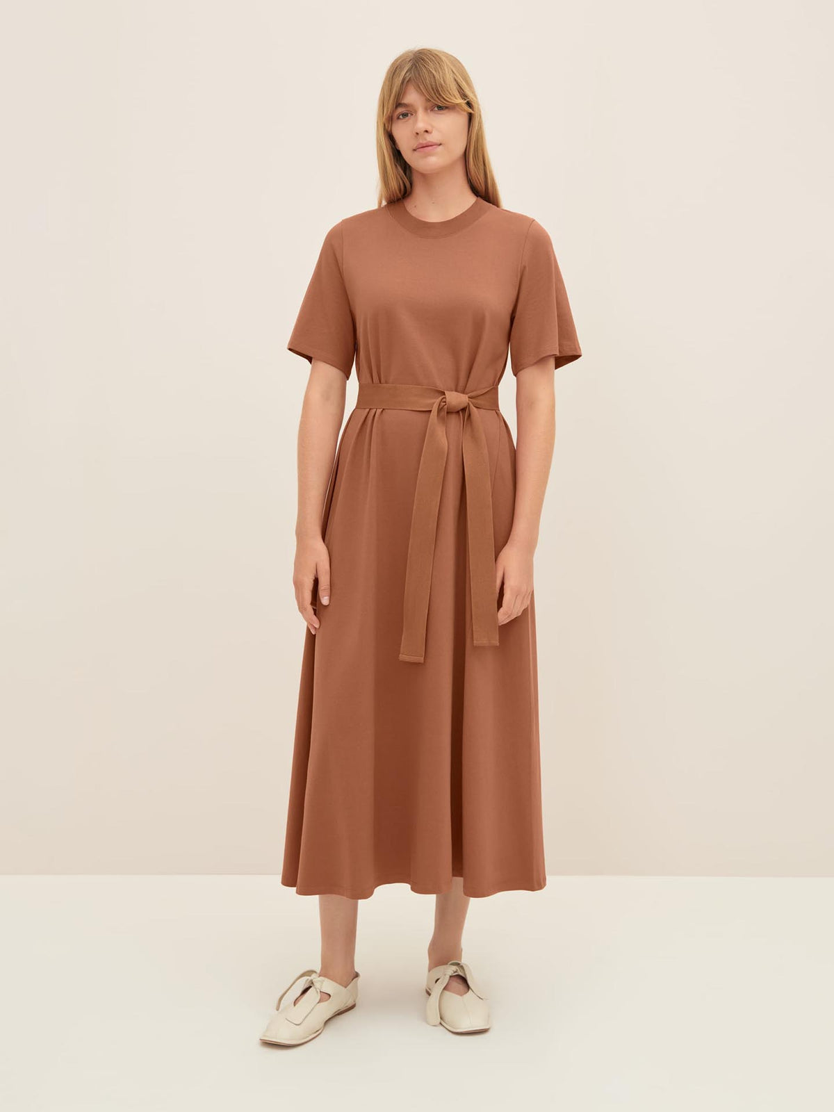 A person wearing Kowtow&#39;s T-Shirt Swing Dress in Sienna and white shoes, standing against a plain background. The short-sleeved, belted midi dress showcases Kowtow&#39;s standard fit beautifully, perfect for those who appreciate both style and comfort.